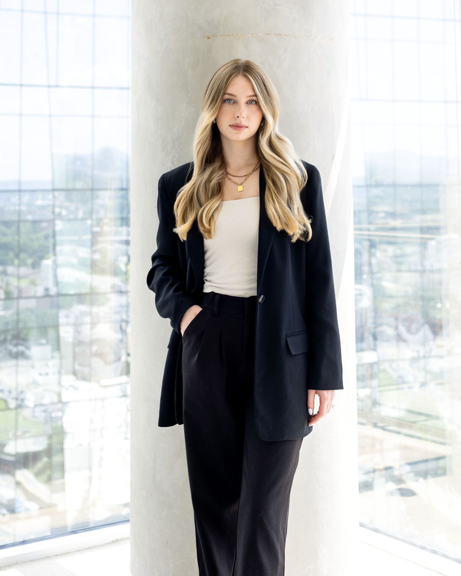 Professional model in a black blazer and white top poses against a modern white interior with city views.