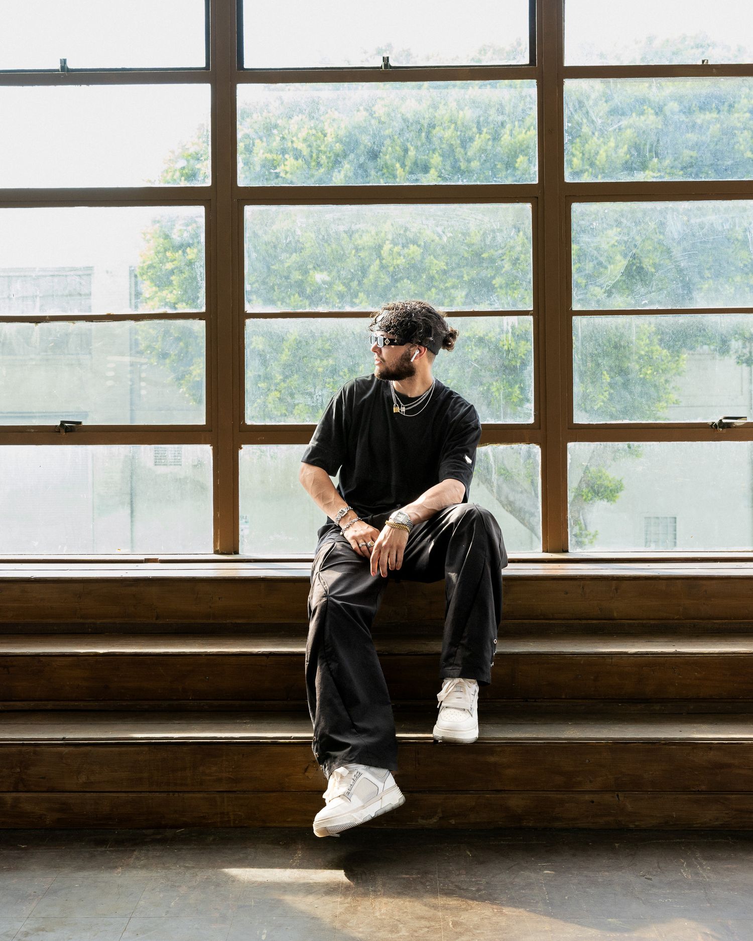 Figure seated alone on wooden windowsill ledge wearing casual black attire and white sneakers in sunlit interior space.