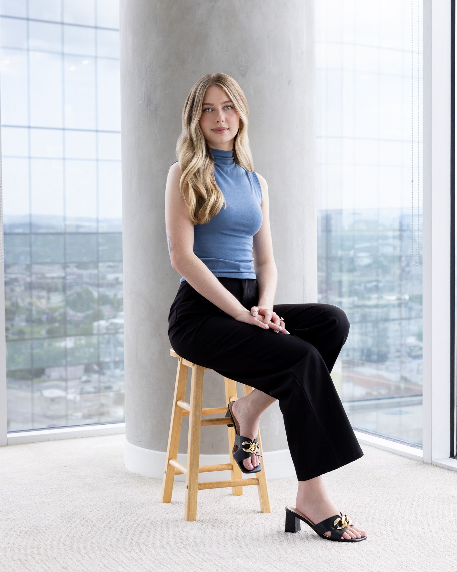 A woman in a light blue top and black pants poses elegantly on wooden stools in a bright, modern space with city views.