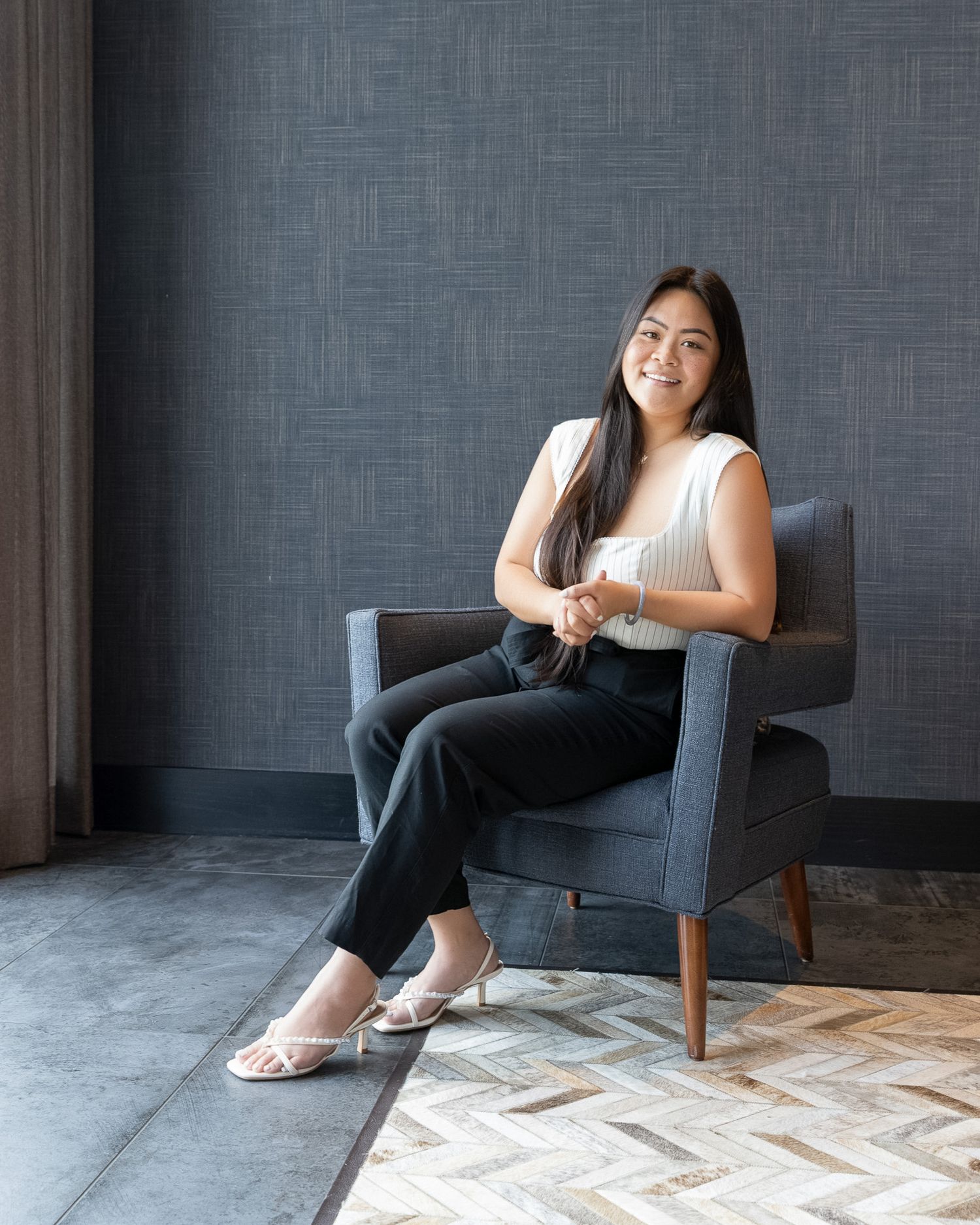 Woman sits relaxed in modern gray armchair against textured wall with pattern on hardwood floor.
