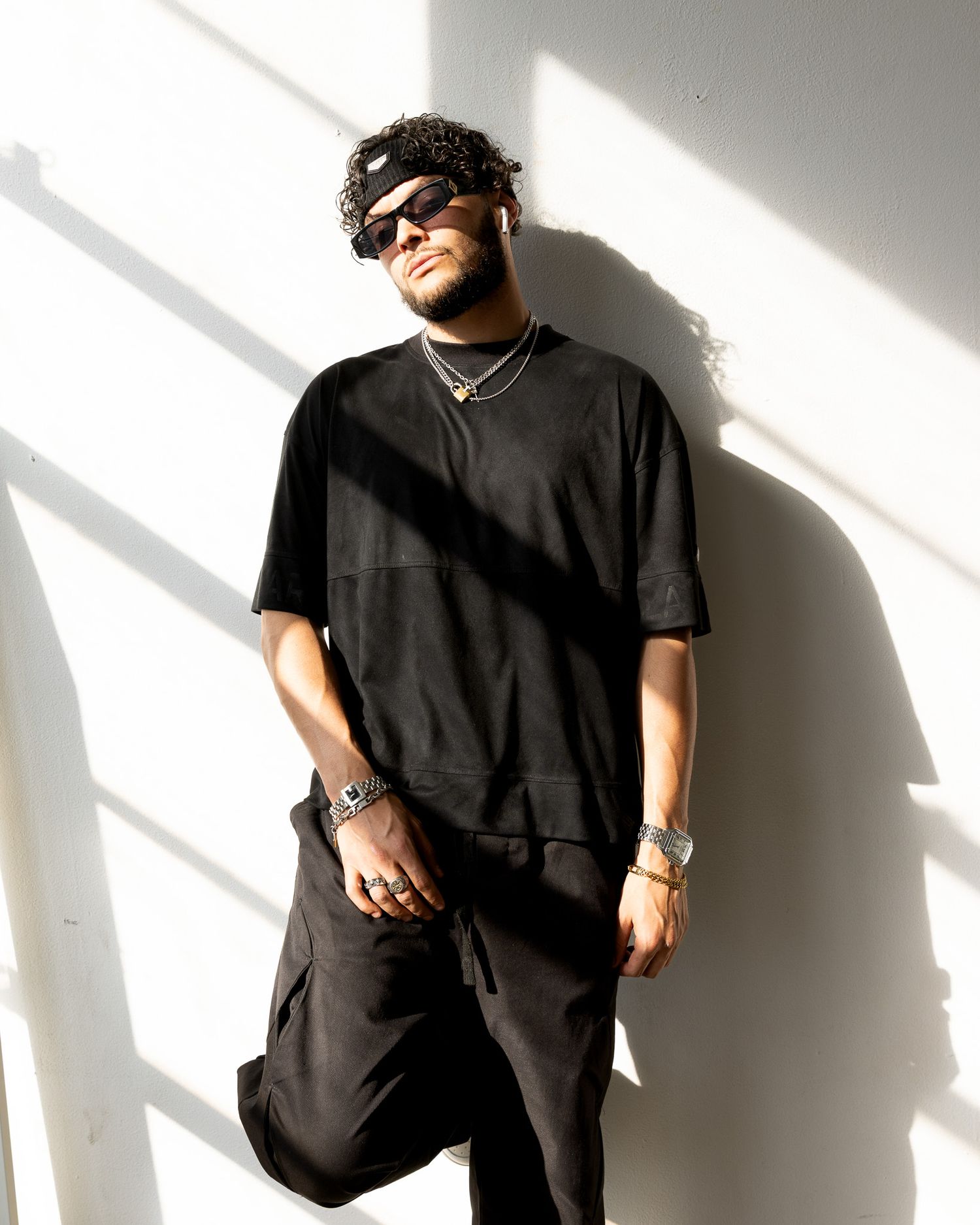Stylish person wearing all black streetwear with sunglasses poses against white wall with dramatic lighting and shadows.