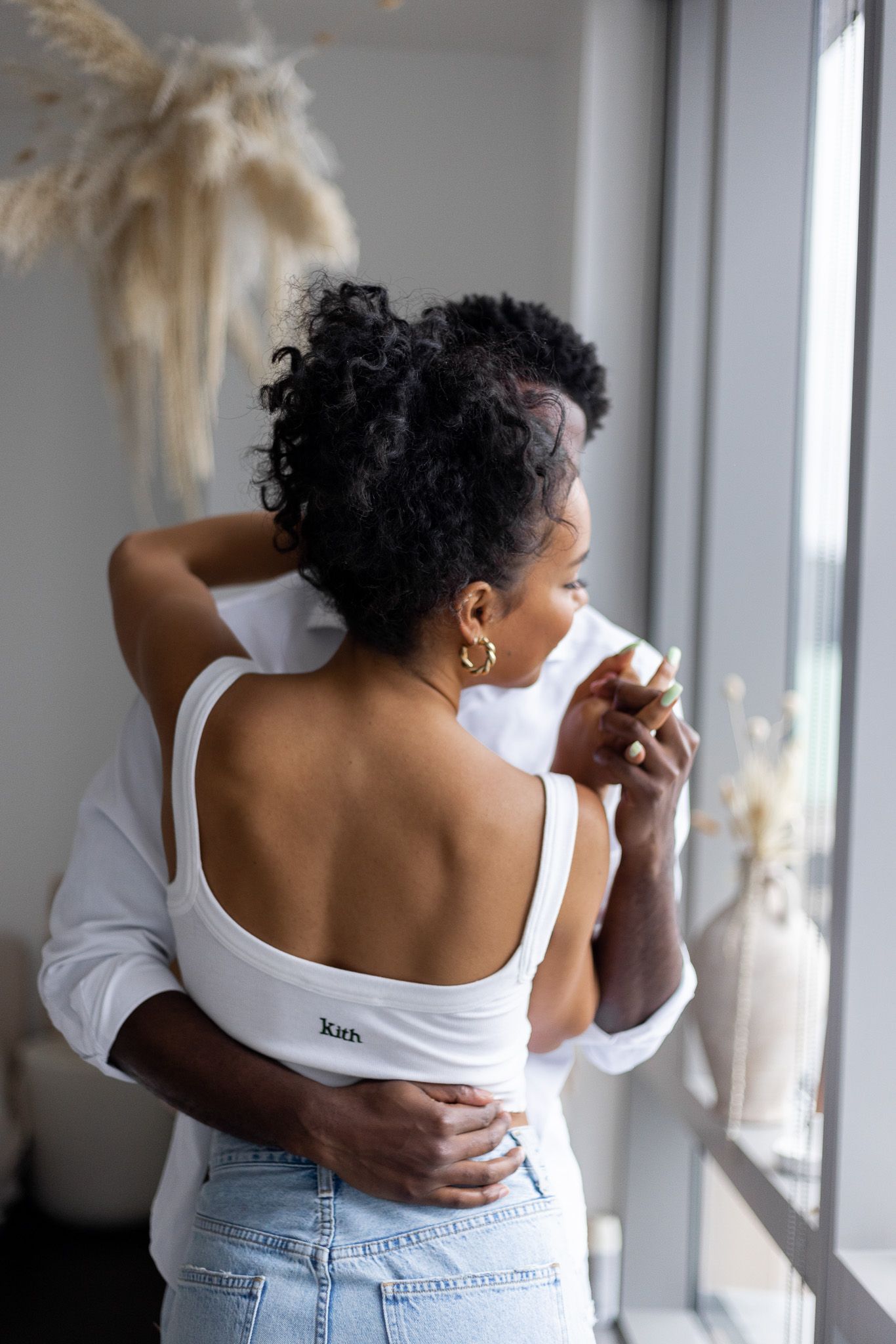 Back view of a person in denim and white top standing near window with curly dark hair and natural lighting.