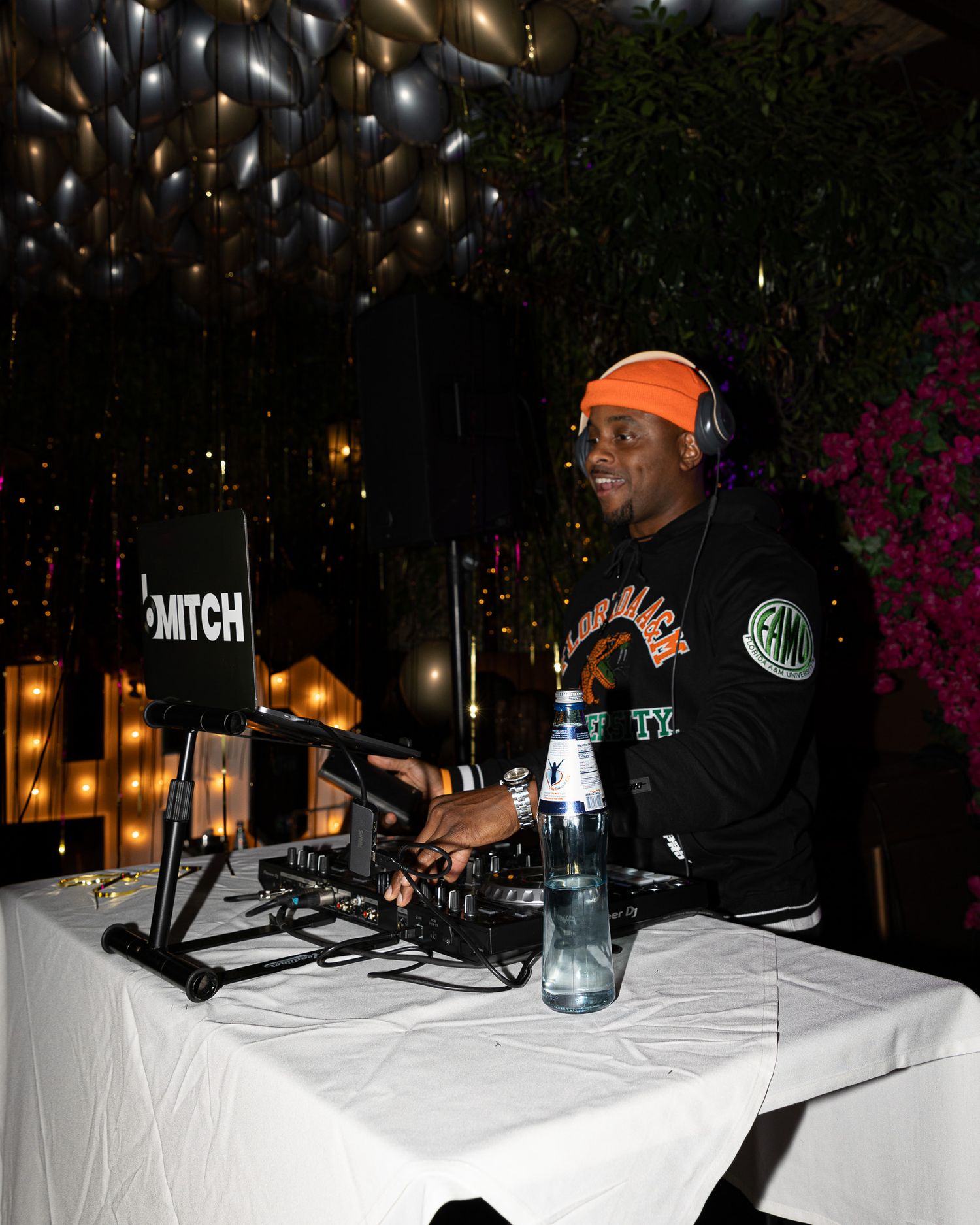 DJ wearing an orange beanie and black sweatshirt performs at a nightclub with Twitch signage and string lights in the background.