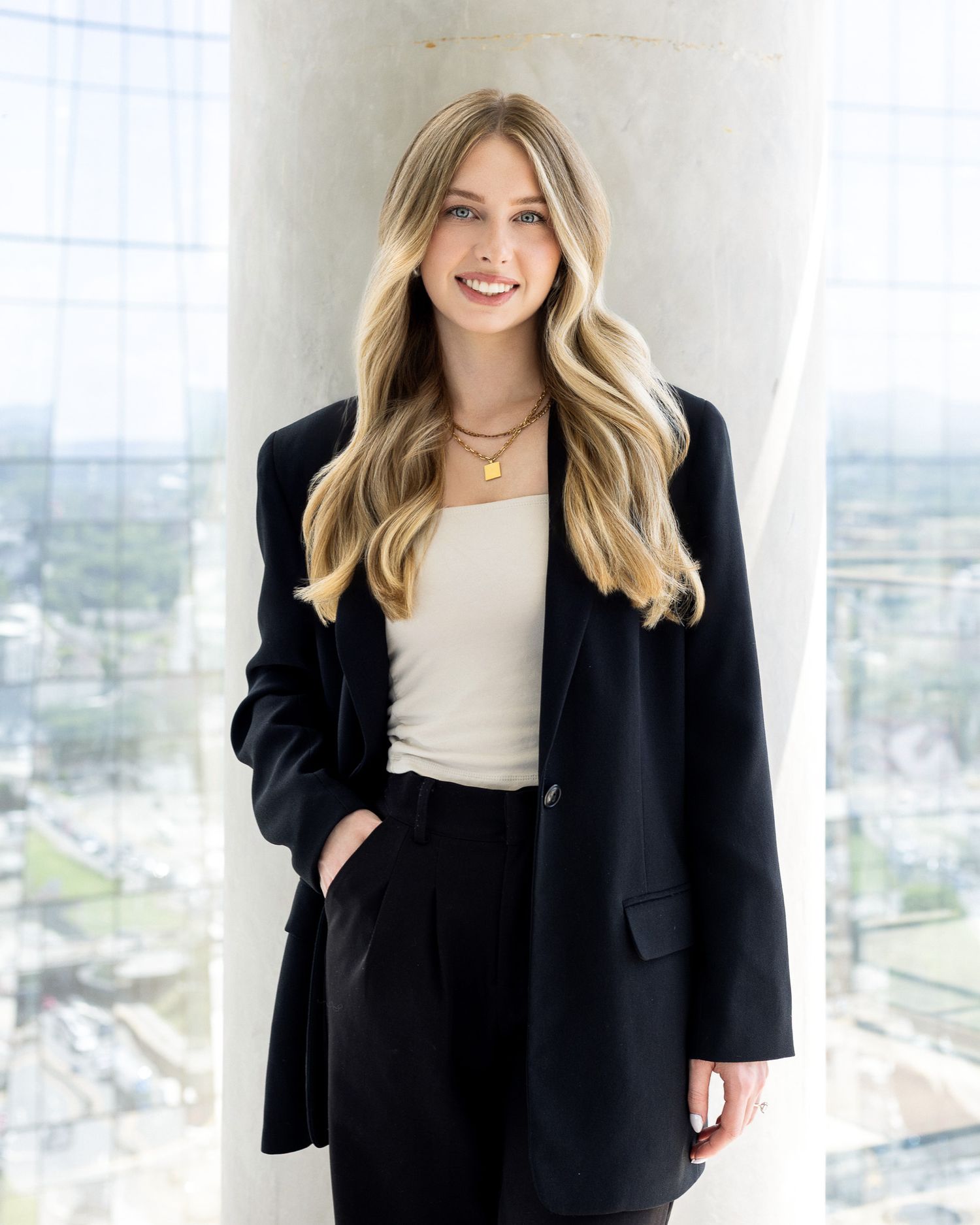 Woman in black blazer and cream top standing in modern office setting with bright windows and city views below.