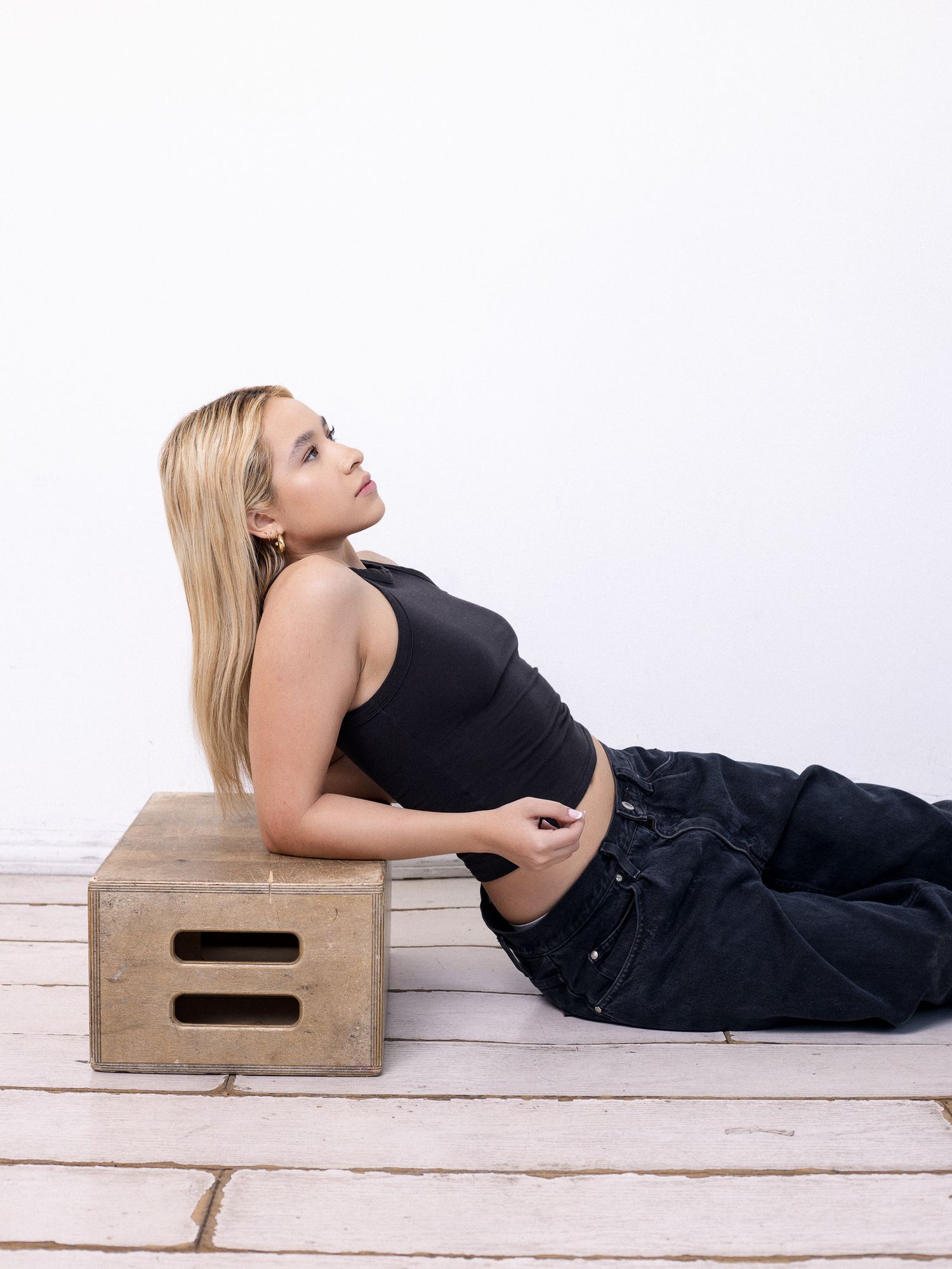 Fashion model wearing black crop top and cargo pants poses while reclining on wooden crate against white backdrop.