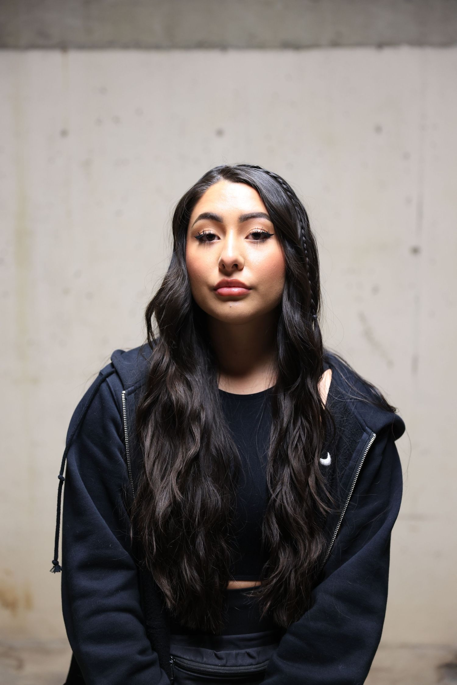 Athletic woman in dark hoodie poses against concrete wall with long black hair.