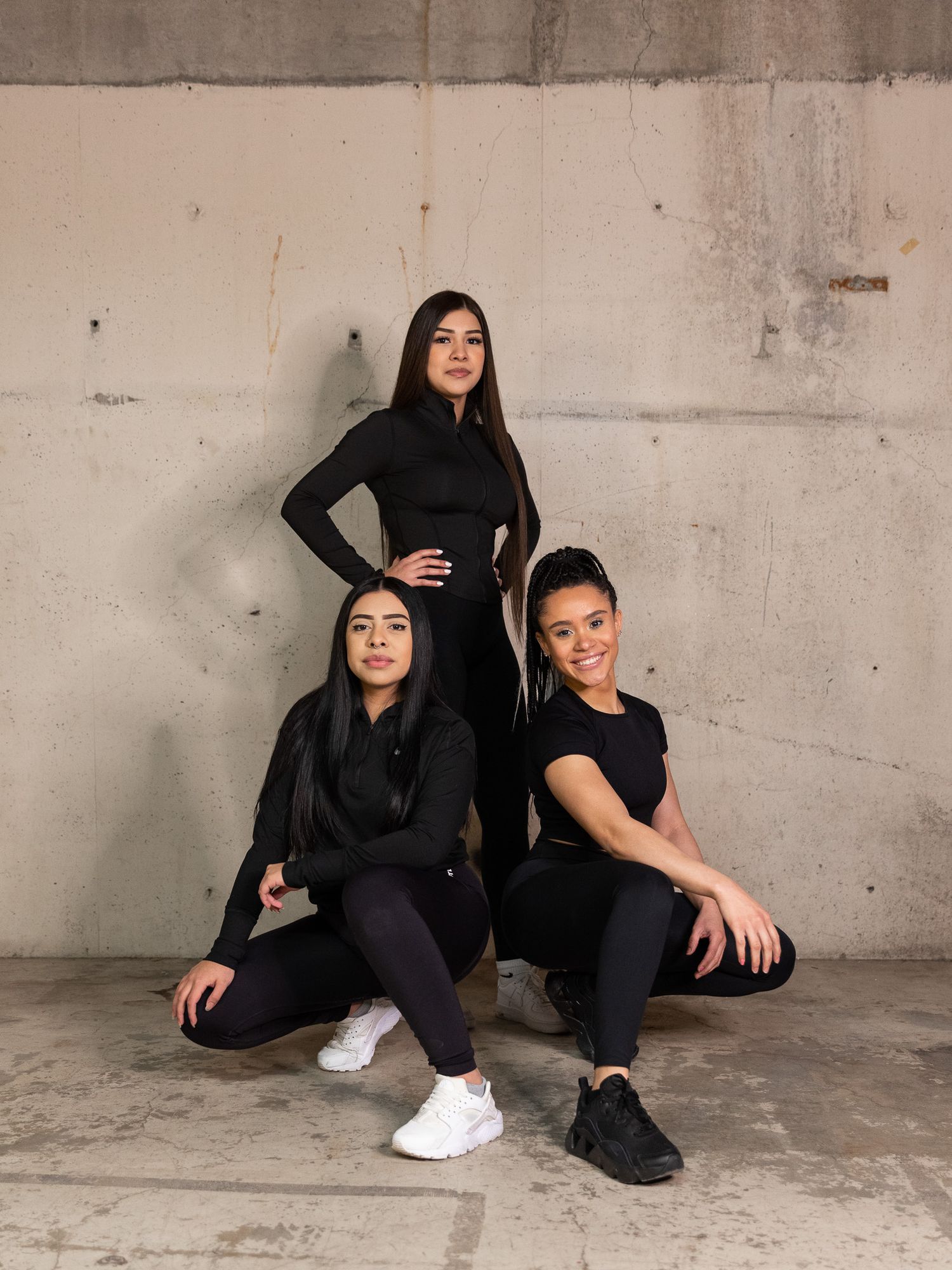 Three women in black athletic wear posing against concrete wall.