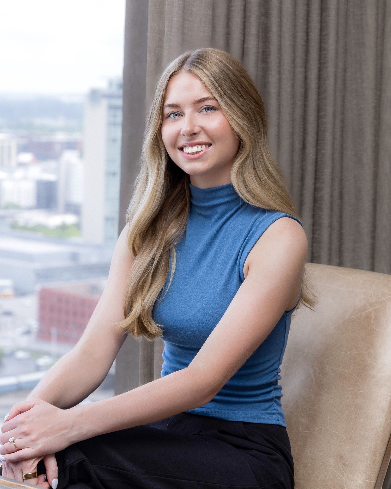 Professional portrait in blue sleeveless turtleneck against urban backdrop through window with warm smile and blonde hair.