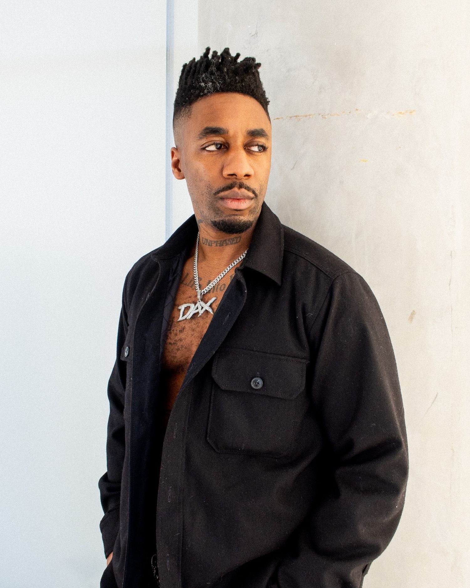 Portrait of person in black button-up jacket with silver chain necklace against light colored wall.