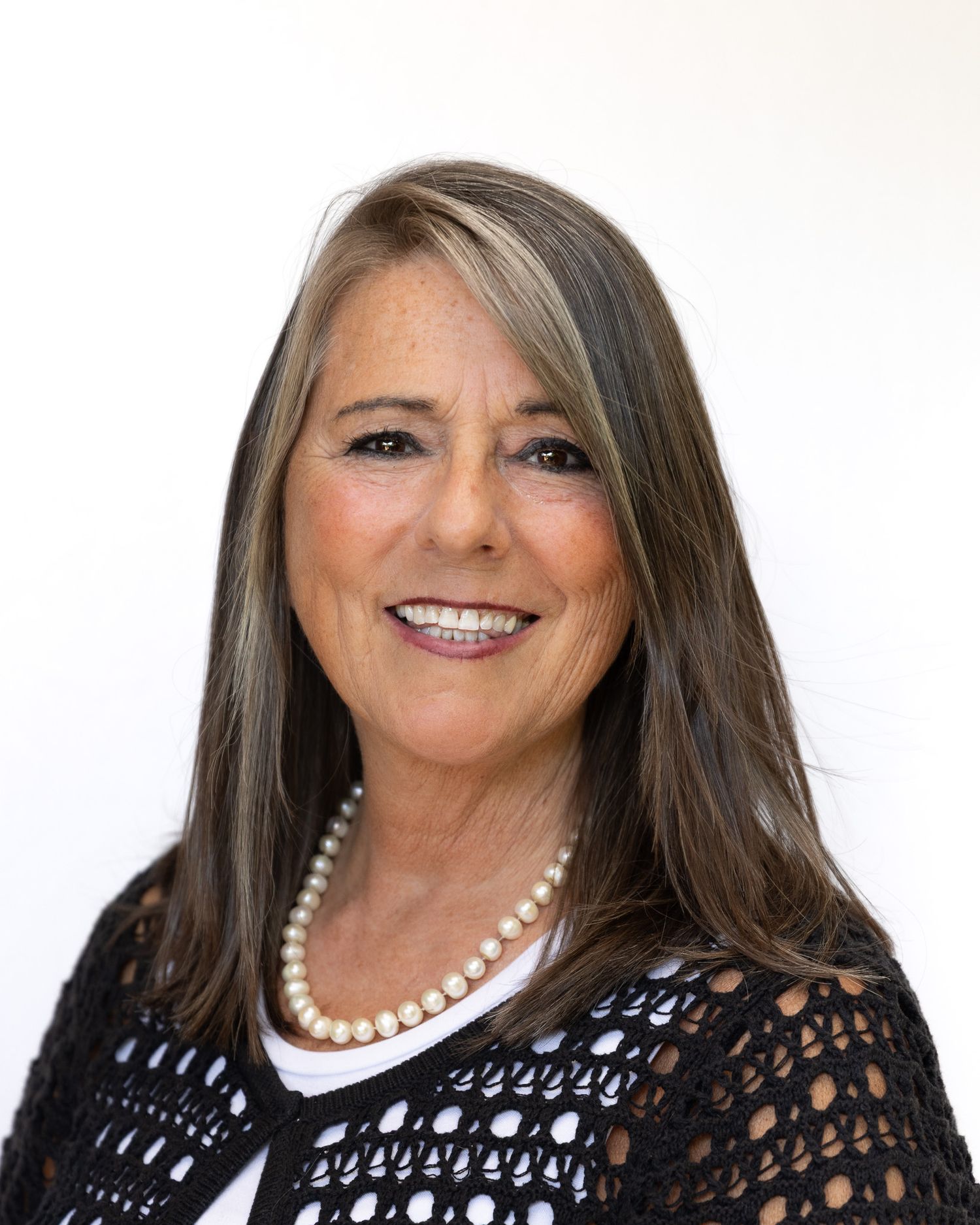 Professional headshot of a person with long brown hair wearing a black lace top and pearl necklace against a white background.