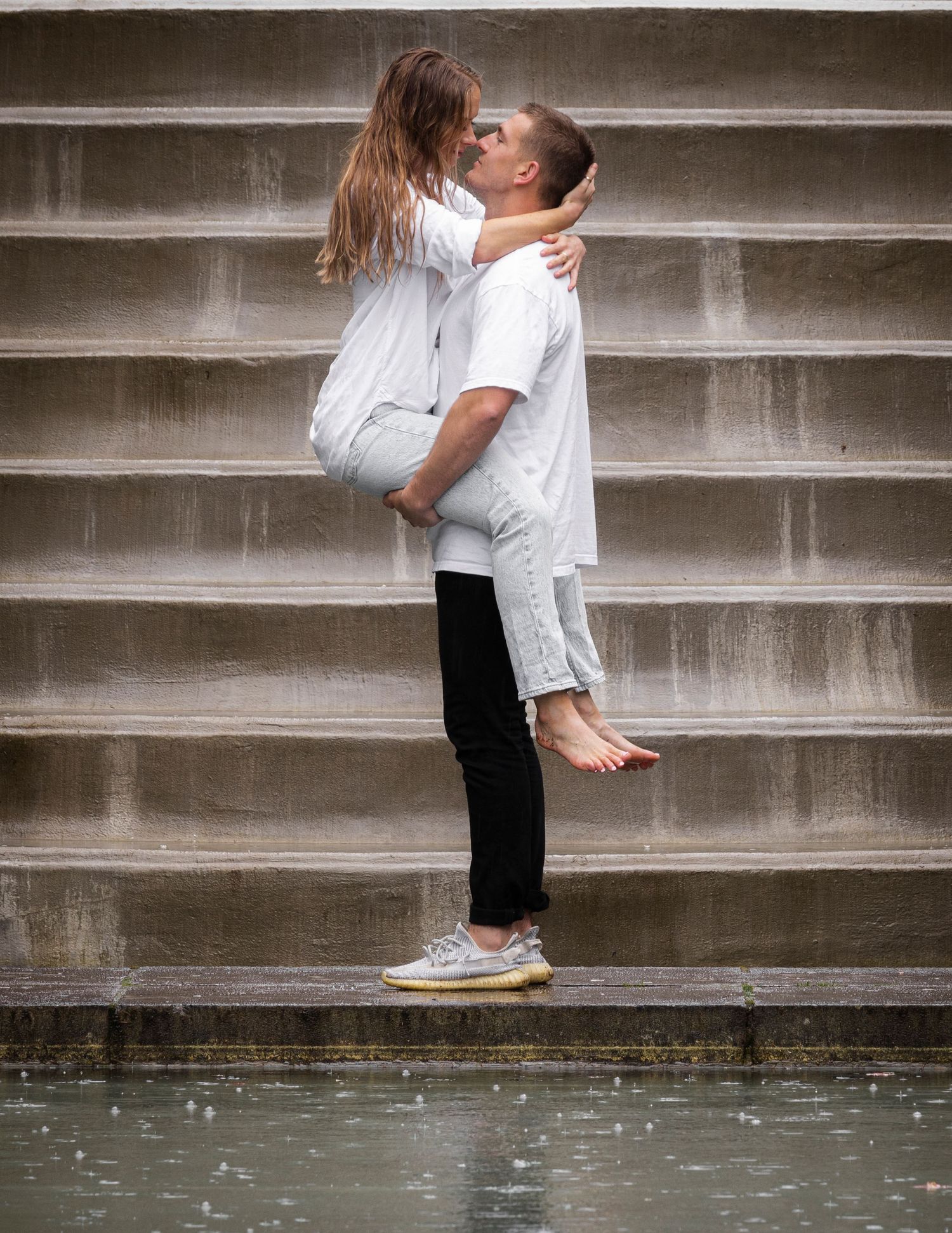 Two people in white outfits share a romantic moment while standing on concrete steps near water.