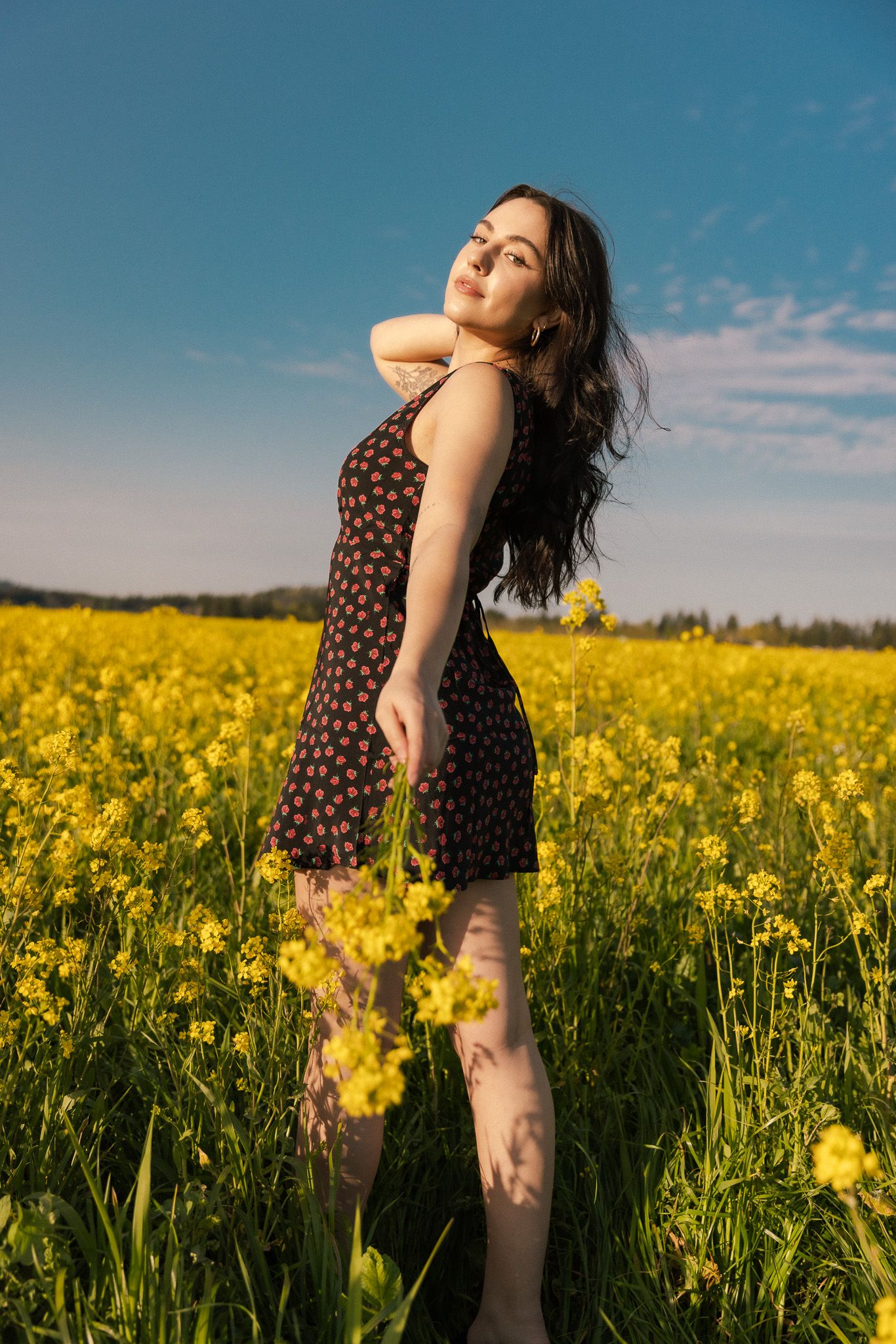 A person in a polka dot dress poses gracefully in a vibrant yellow canola field under bright blue summer skies.