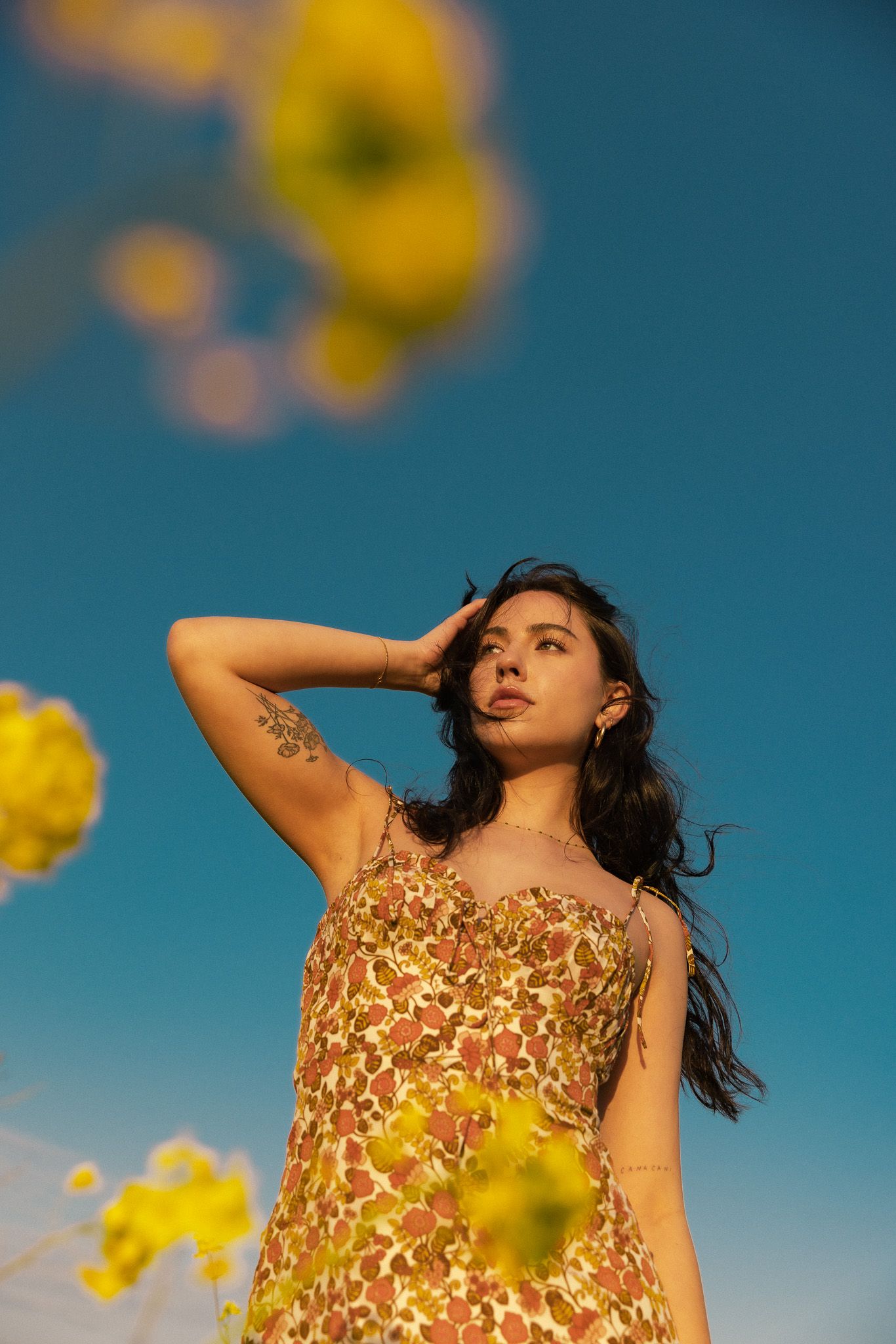 Person in a floral dress poses against blue sky with yellow flowers in soft focus in the foreground.