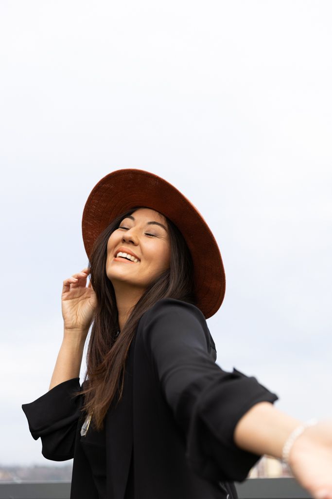 A joyful person wearing a brown hat and black top spins playfully against a bright sky background.