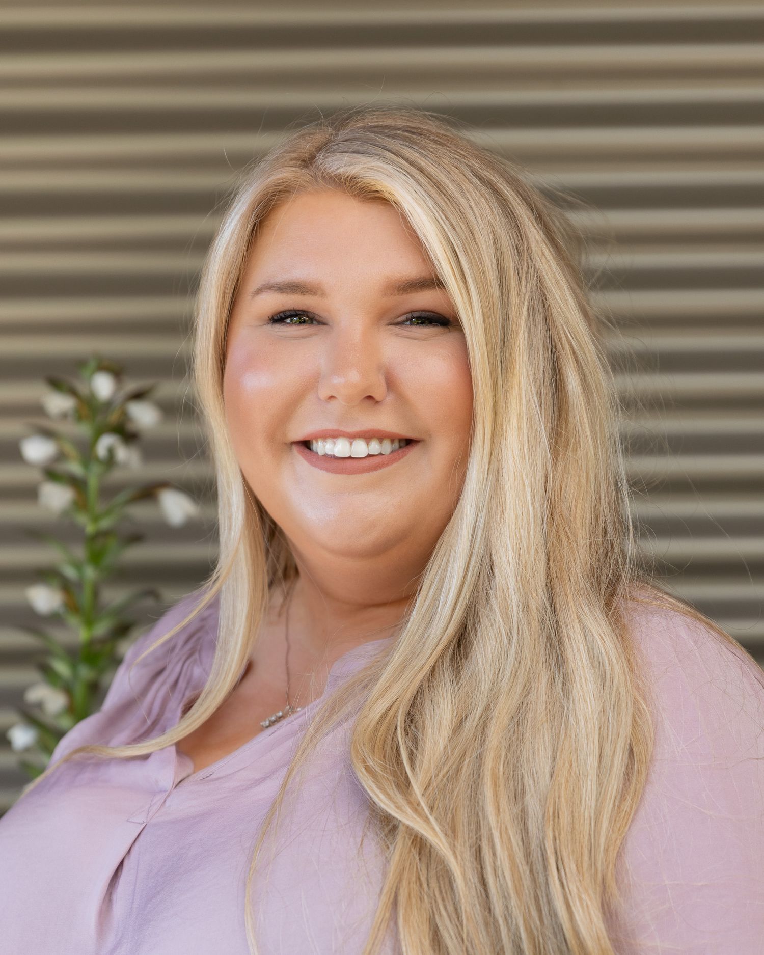 Professional headshot of a person in a light pink top with long blonde hair and a warm smile against a textured background.