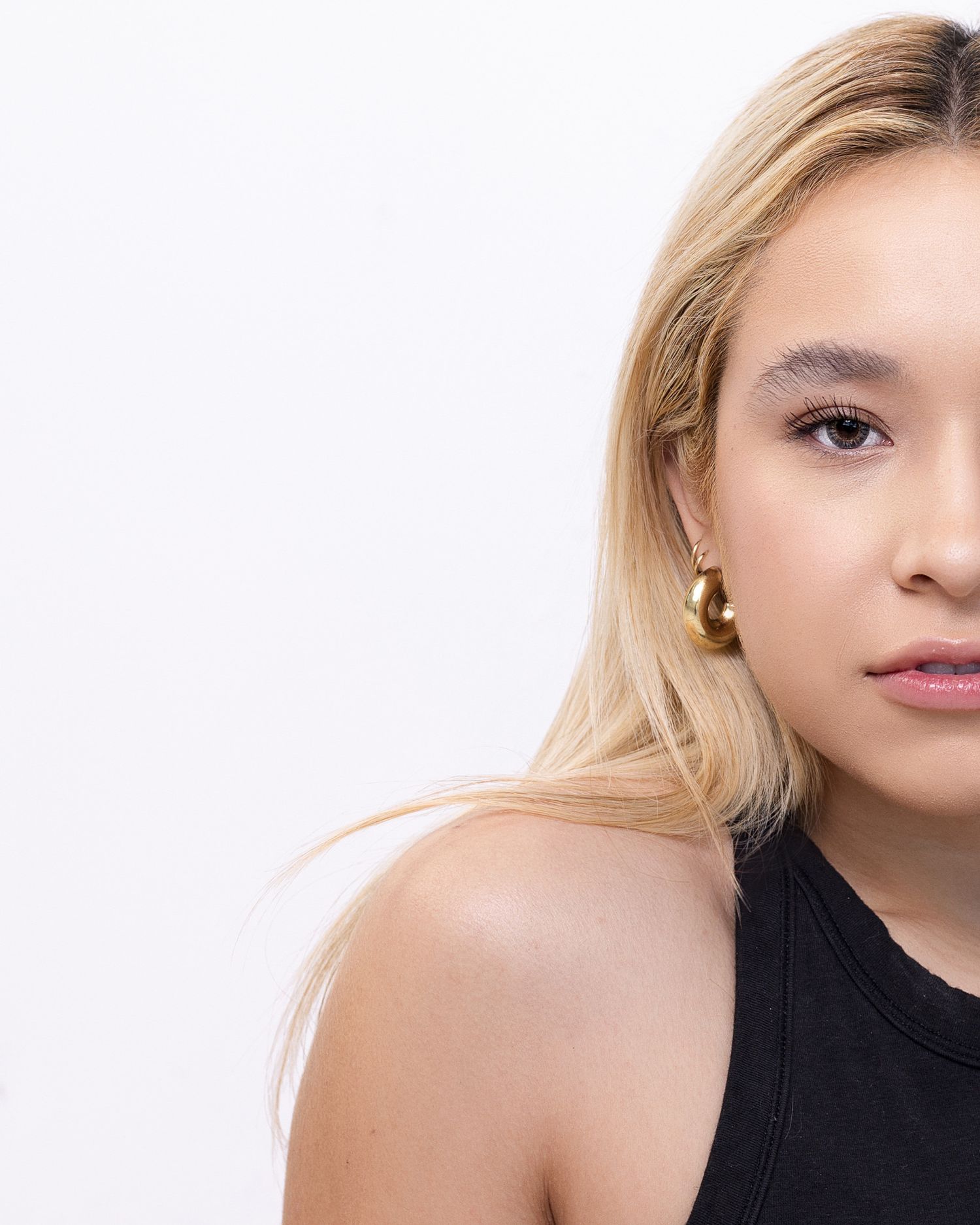 Stylish portrait photograph showing blonde hair against white background, wearing black sleeveless top and gold earrings.
