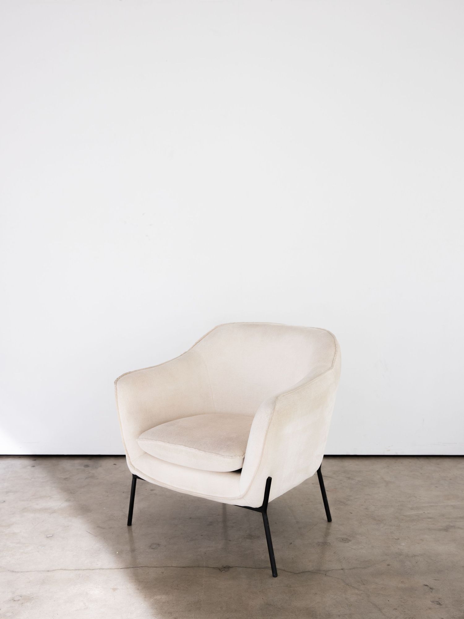 Modern cream-colored velvet armchair with curved back and metal legs sits elegantly against white wall on concrete floor.