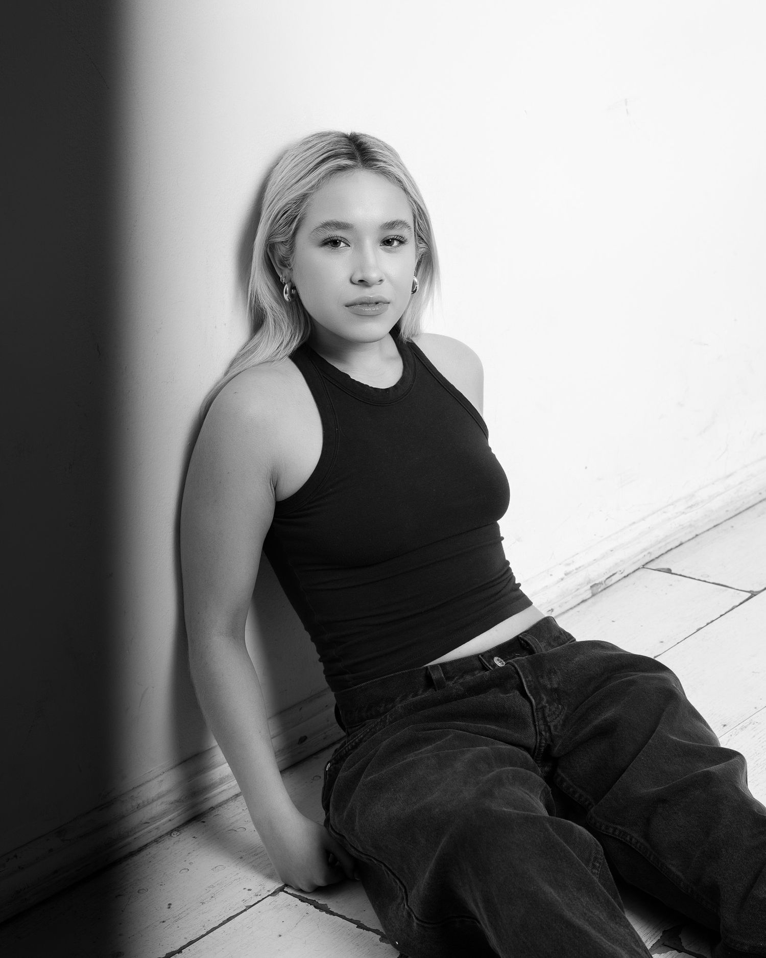 Black and white portrait of someone sitting against a wall wearing a black tank top and dark pants.