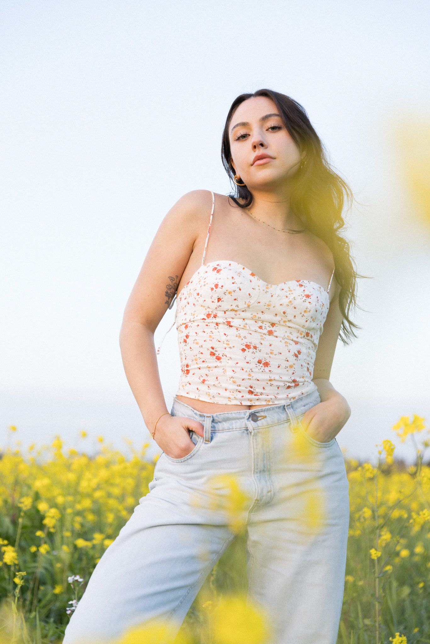 Person in a floral crop top and light jeans poses in a sunny yellow canola field during golden hour.