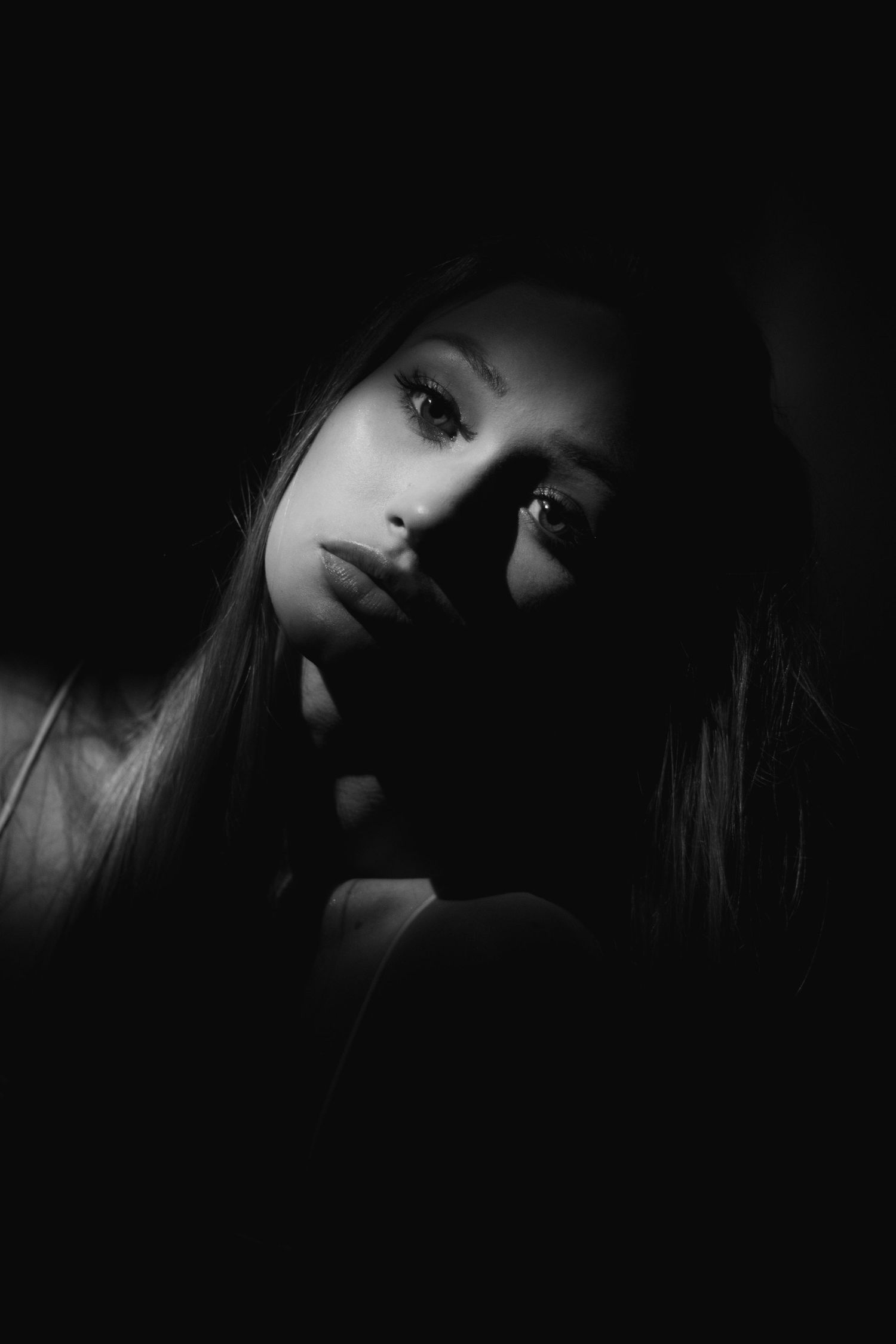 A dramatic black and white portrait with high contrast lighting illuminating one side of a face emerging from darkness.