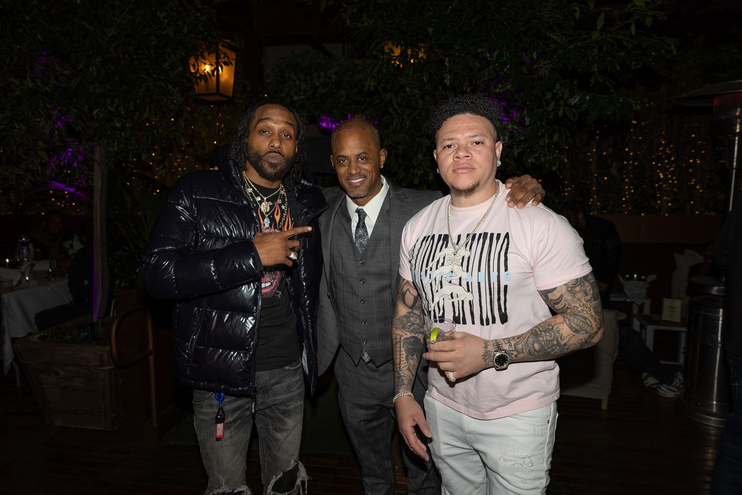 Three people pose together at a nighttime event with one wearing a dark puffy jacket and another in a business suit.