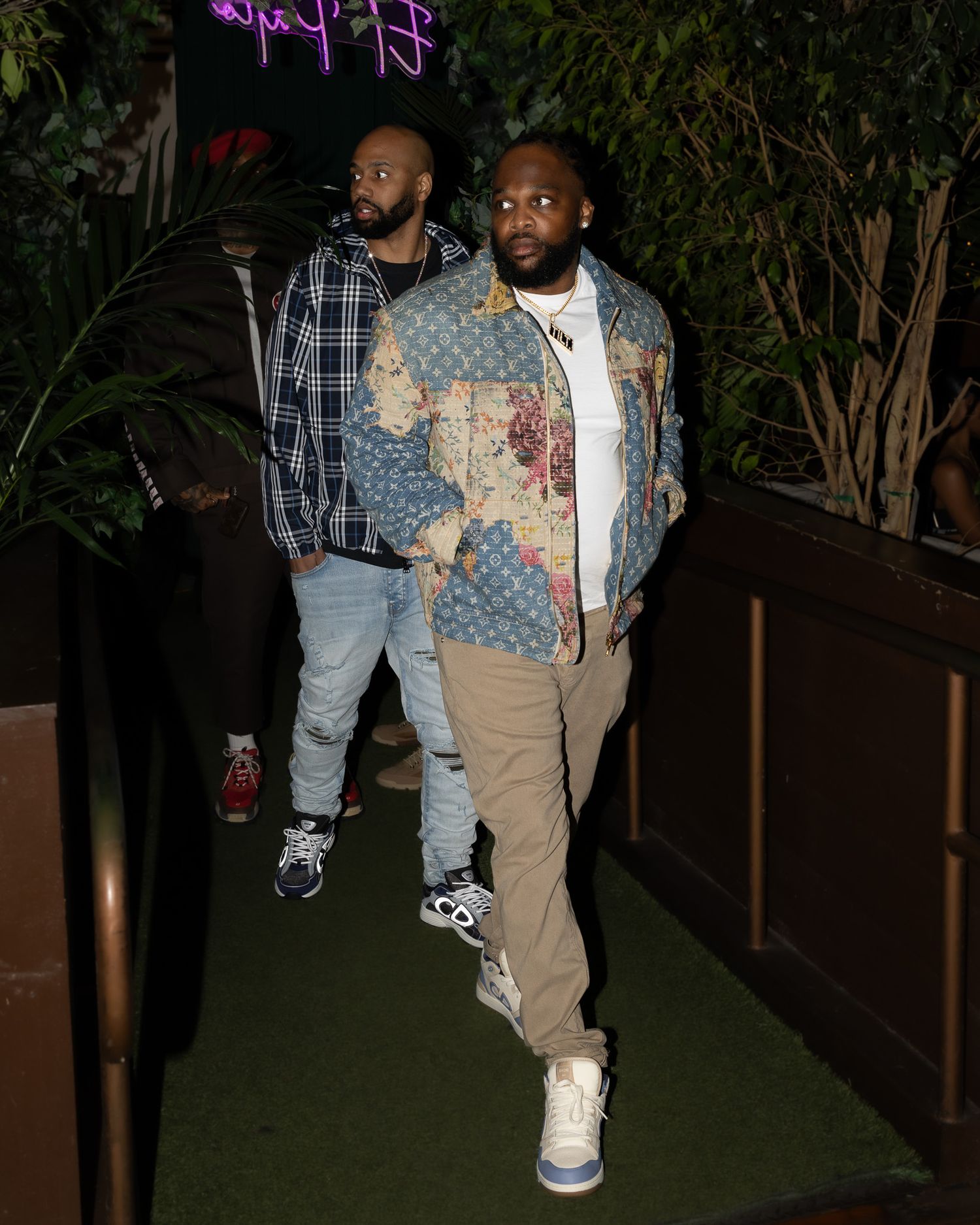 Two men walking on a green-carpeted pathway at night wearing casual streetwear including plaid and denim jackets.