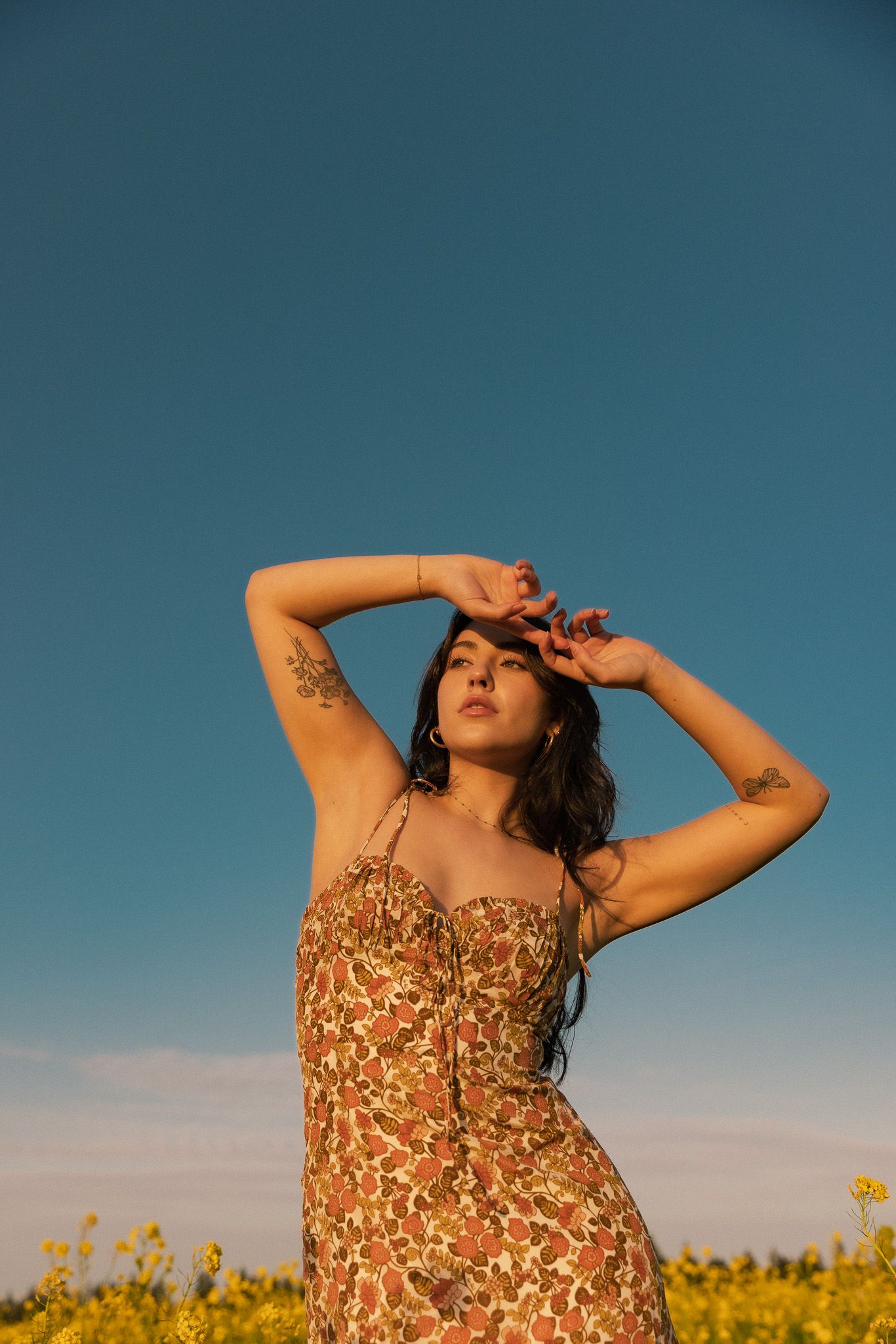 A woman in a floral dress poses in a golden field at sunset with her arms raised against a deep blue sky.