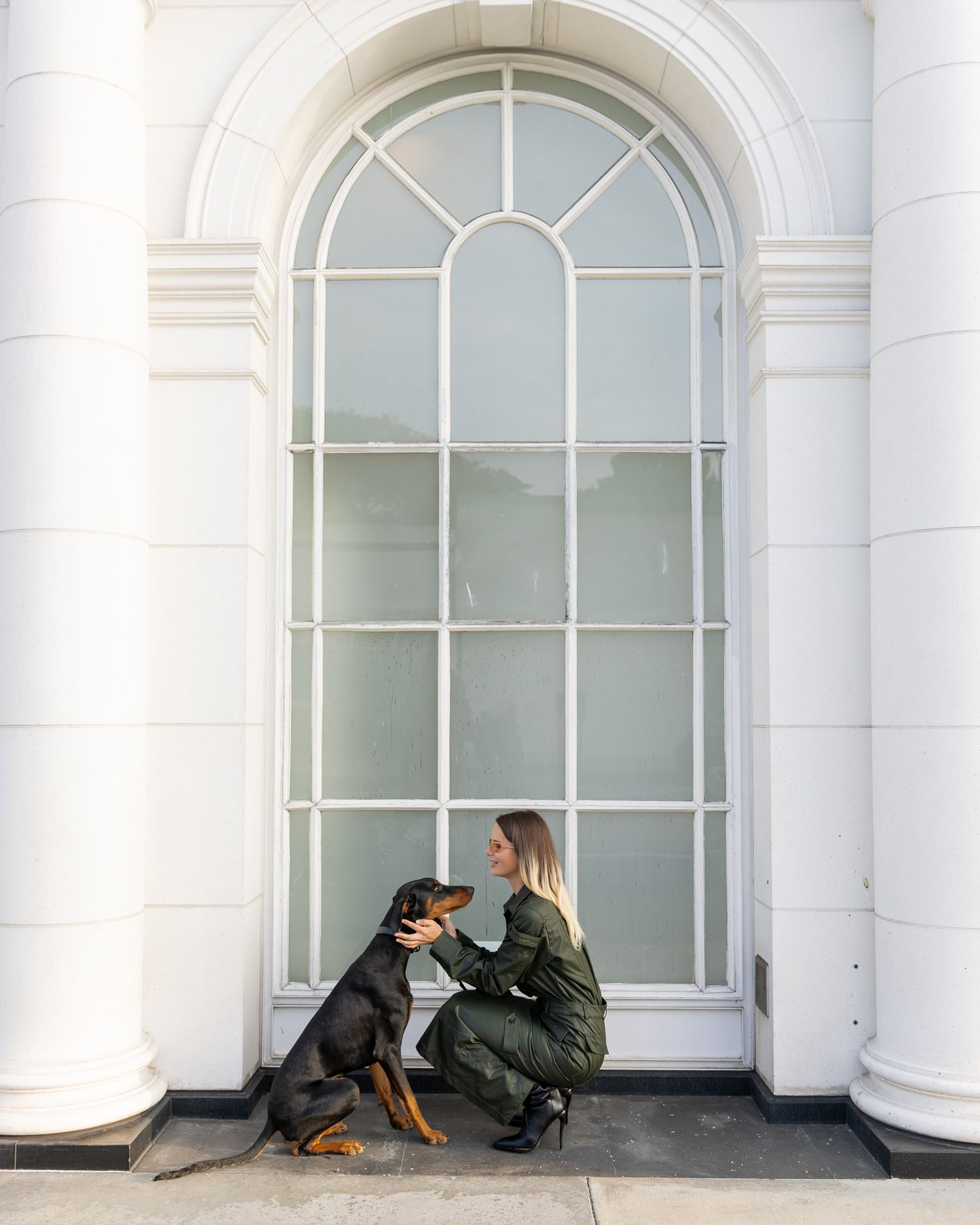 A person in a green jacket crouches next to a black and brown dog by a tall arched window of a white building.