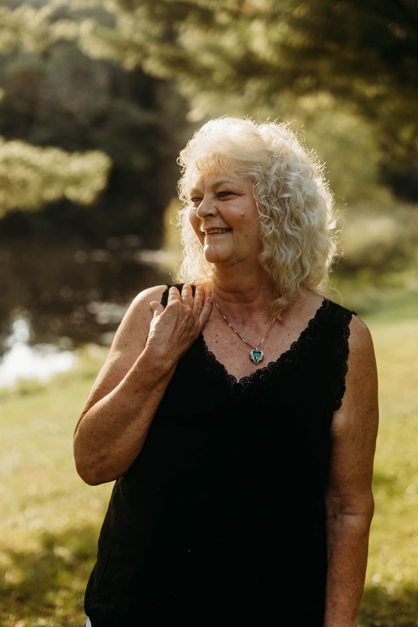 A mature woman in a black dress smiles warmly in natural outdoor lighting.