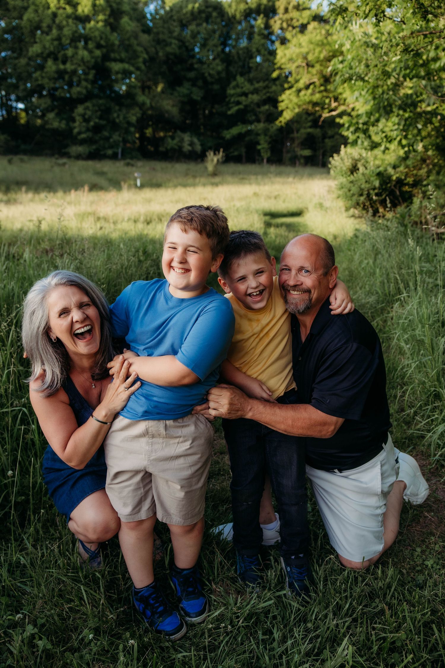 Happy family of four laughing together outdoors in a lush green field during a warm summer day.