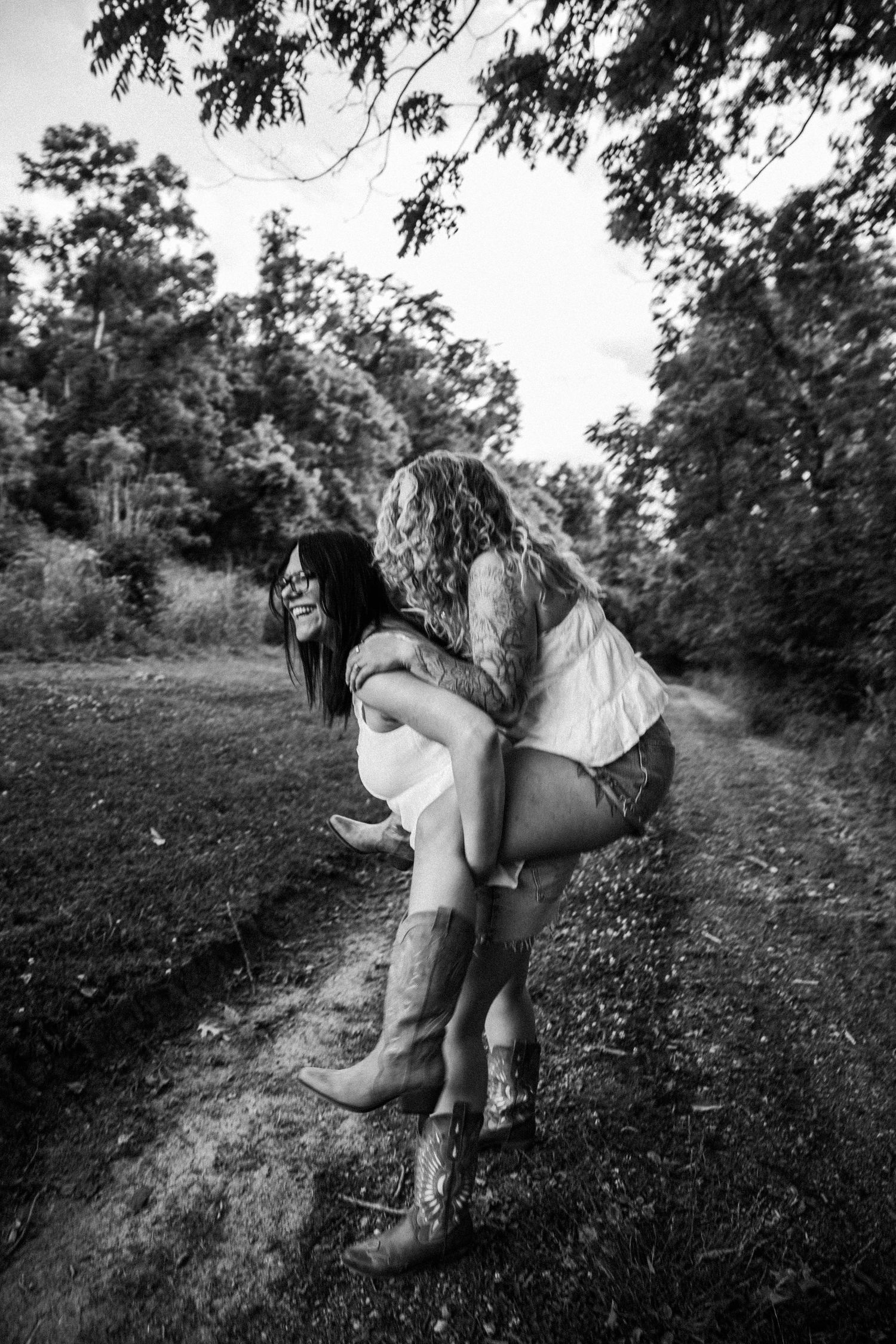 Two young women laughing outdoors, one giving the other a piggyback ride on a dirt path surrounded by trees.