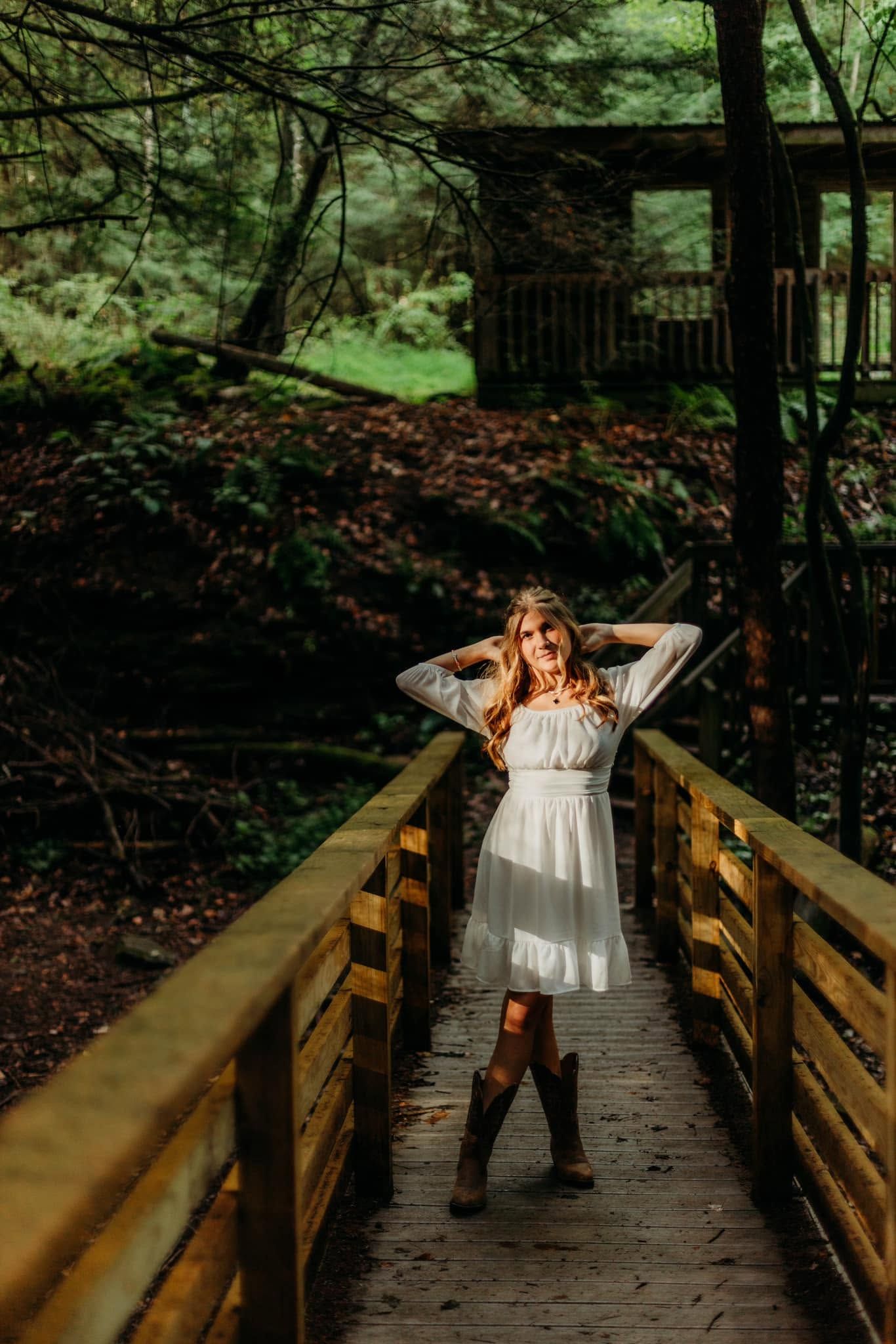 Young woman in white dress and cowboy boots posing on a wooden bridge surrounded by lush forest.