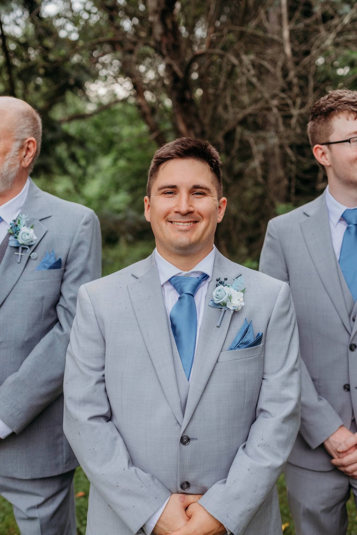 Groomsmen in light gray suits with blue ties and boutonnieres standing outdoors.
