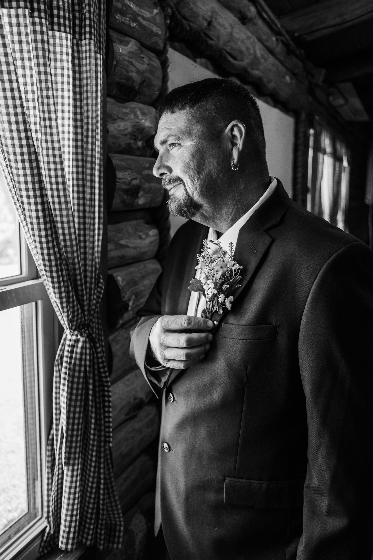 Black and white photo of a groom in a suit with boutonniere gazing out a log cabin window on his wedding day.