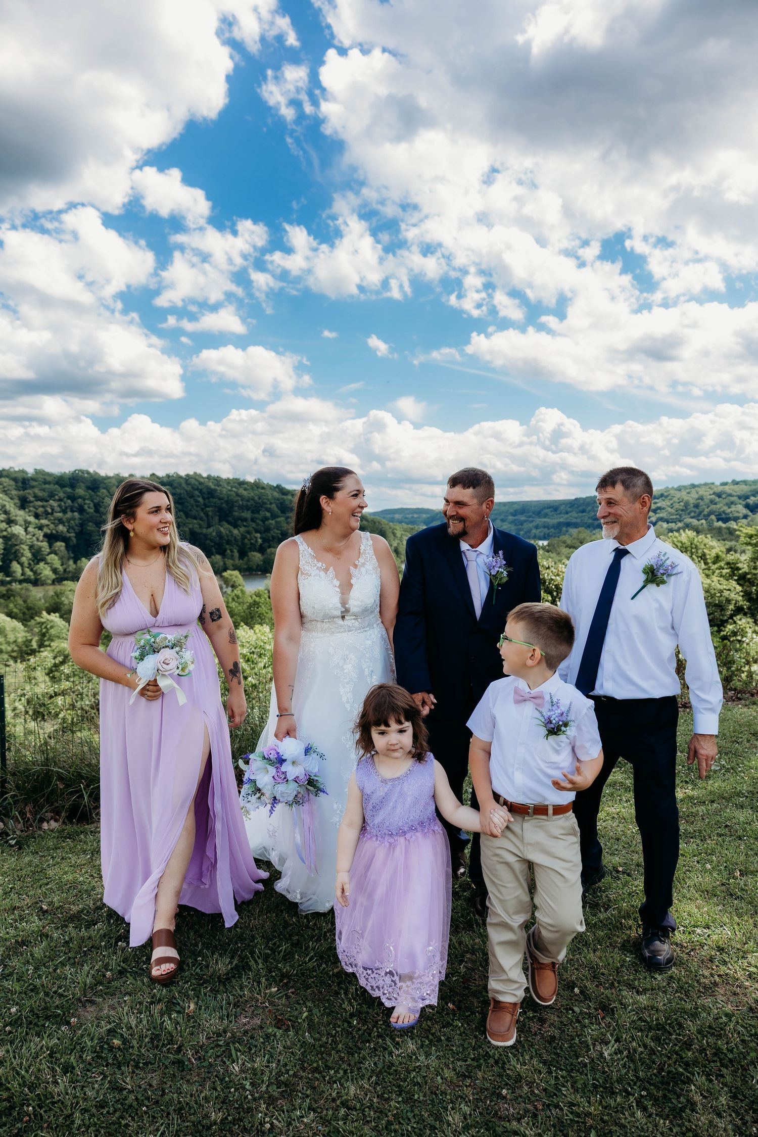 Wedding party walks outdoors, bride in white gown, bridesmaid in lavender, groomsmen in suits, two children in matching outfits.