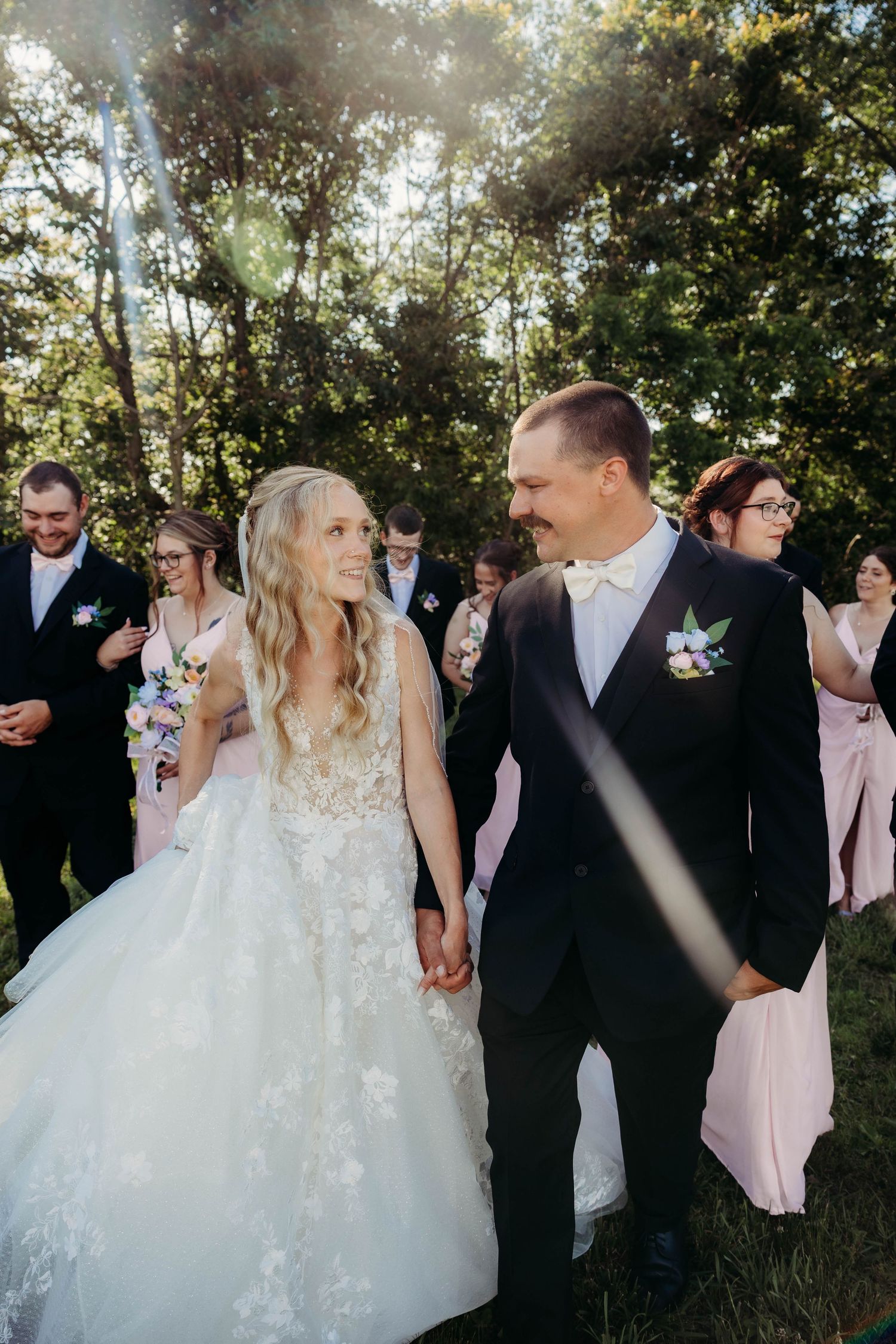 Bride and groom hold hands smiling outdoors with wedding party in background amid sunlit trees.