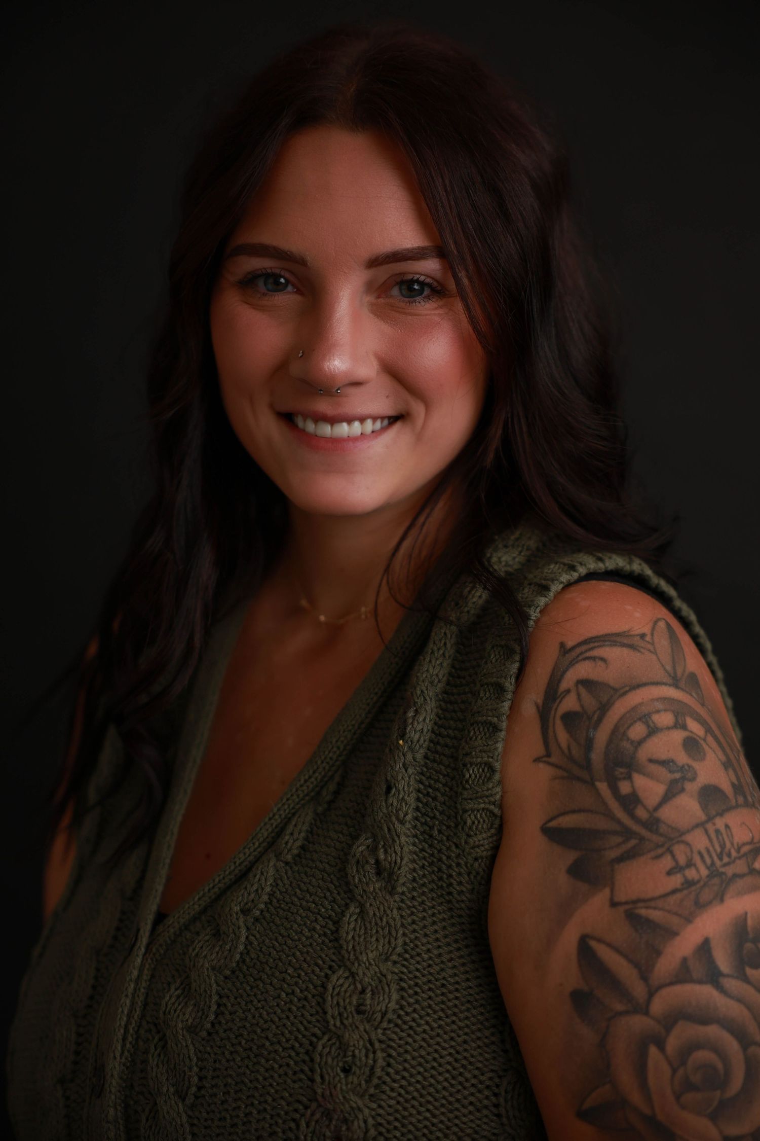 Studio portrait featuring olive green top and artistic arm tattoo against black background.