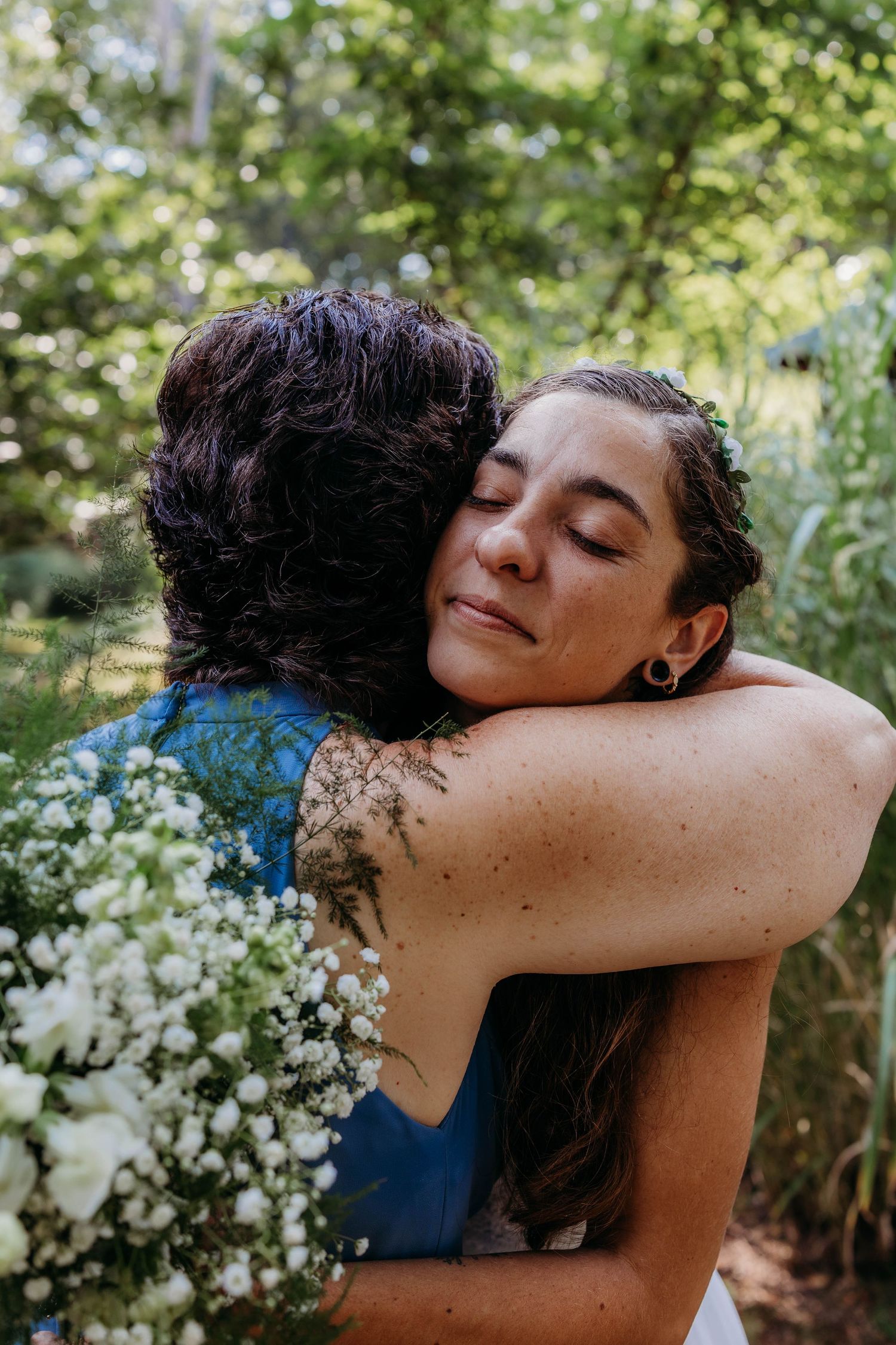 Two people sharing a warm embrace outdoors surrounded by greenery and white flowers.
