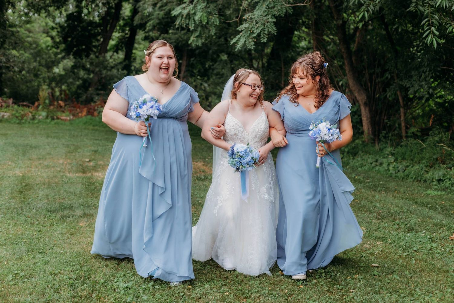 Bridesmaids in light blue dresses walking together with bouquets on grassy lawn.
