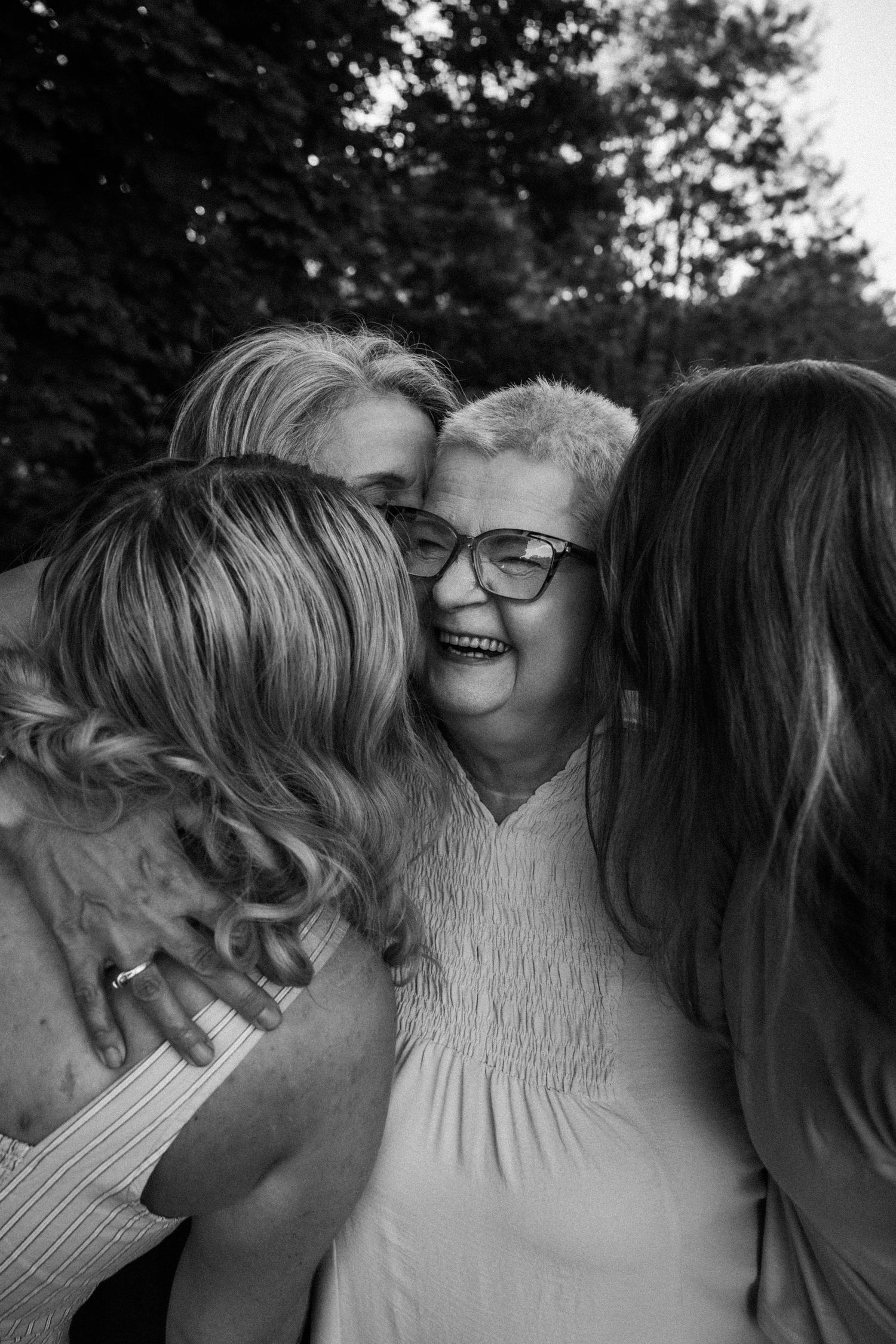 Black and white photo of an older woman with glasses smiling warmly while being hugged by two younger women outdoors.