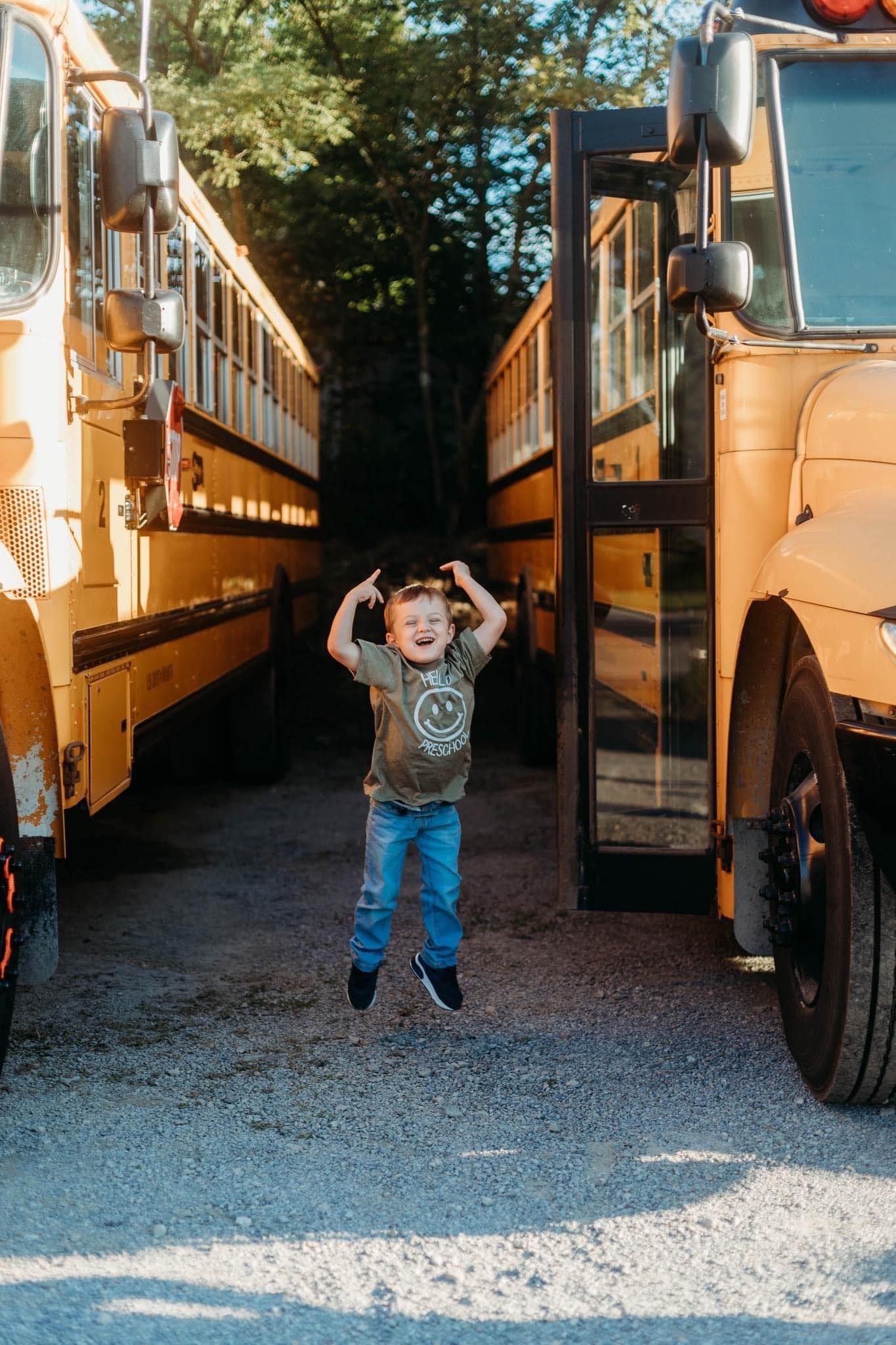 Small child jumps playfully between two parked yellow school buses on a sunny day.