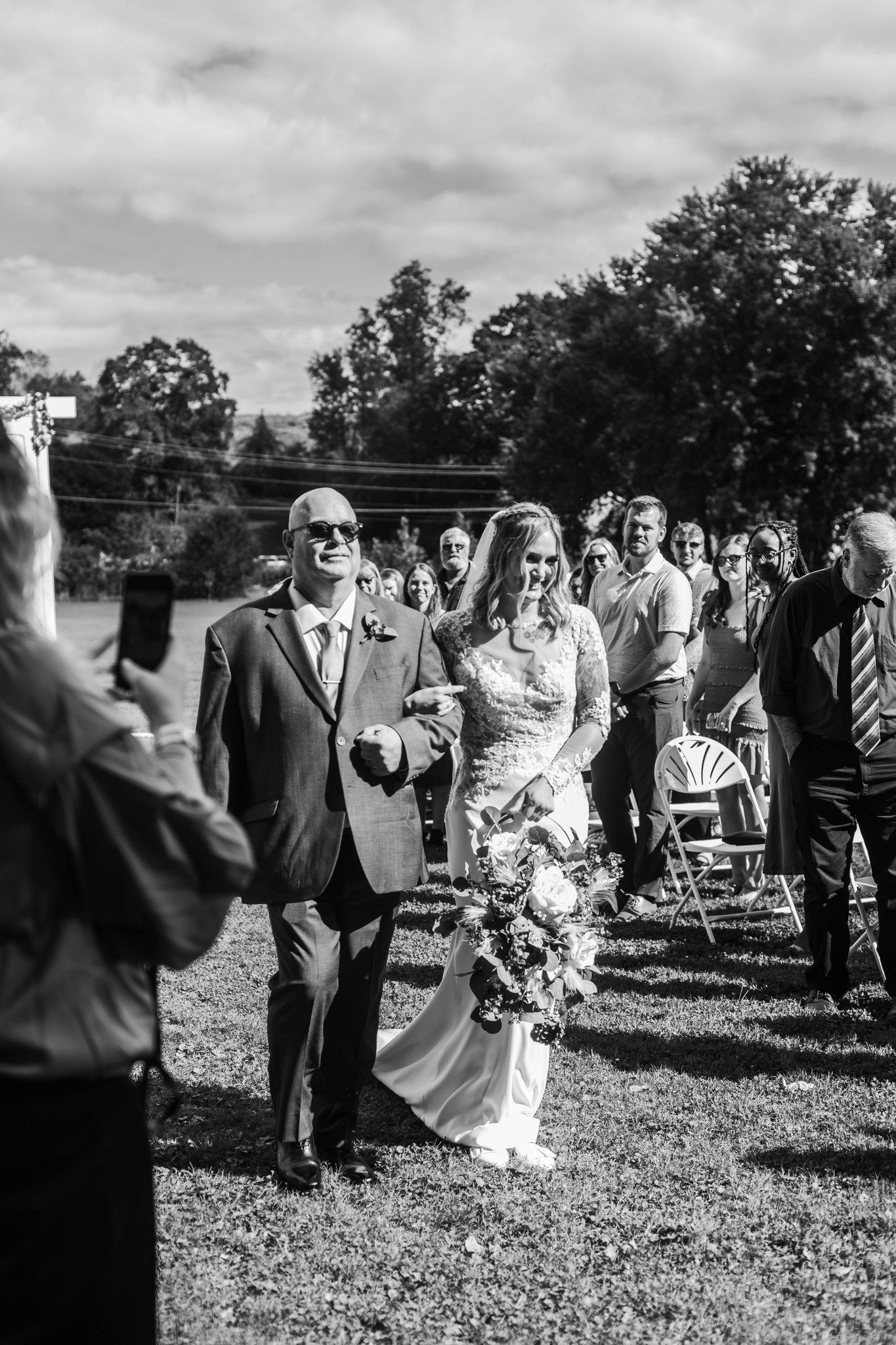 A black and white photo of a wedding ceremony processional on a grassy lawn with trees in the background.