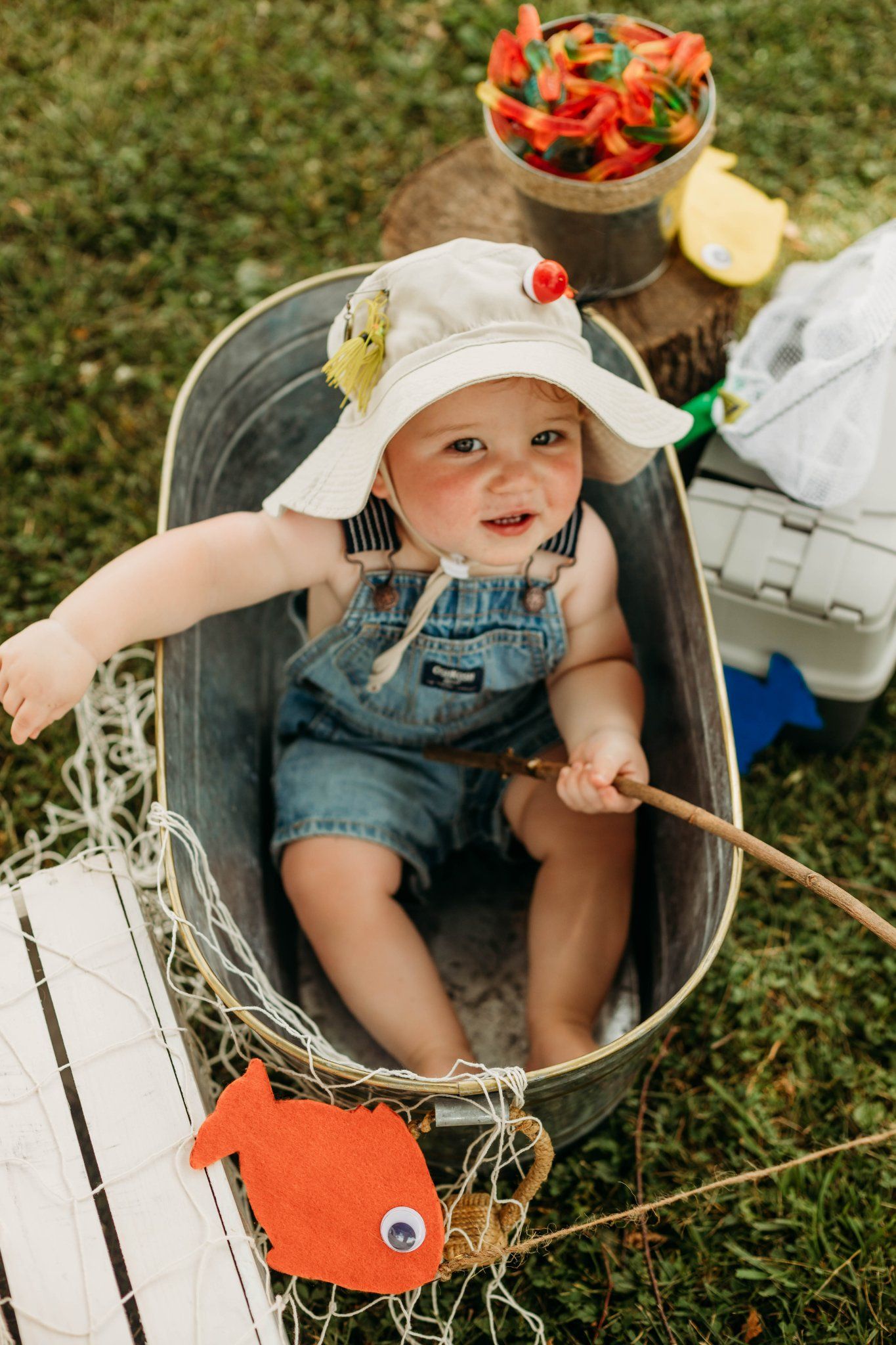 Baby in overalls and sun hat sitting in metal tub with fishing-themed decorations.