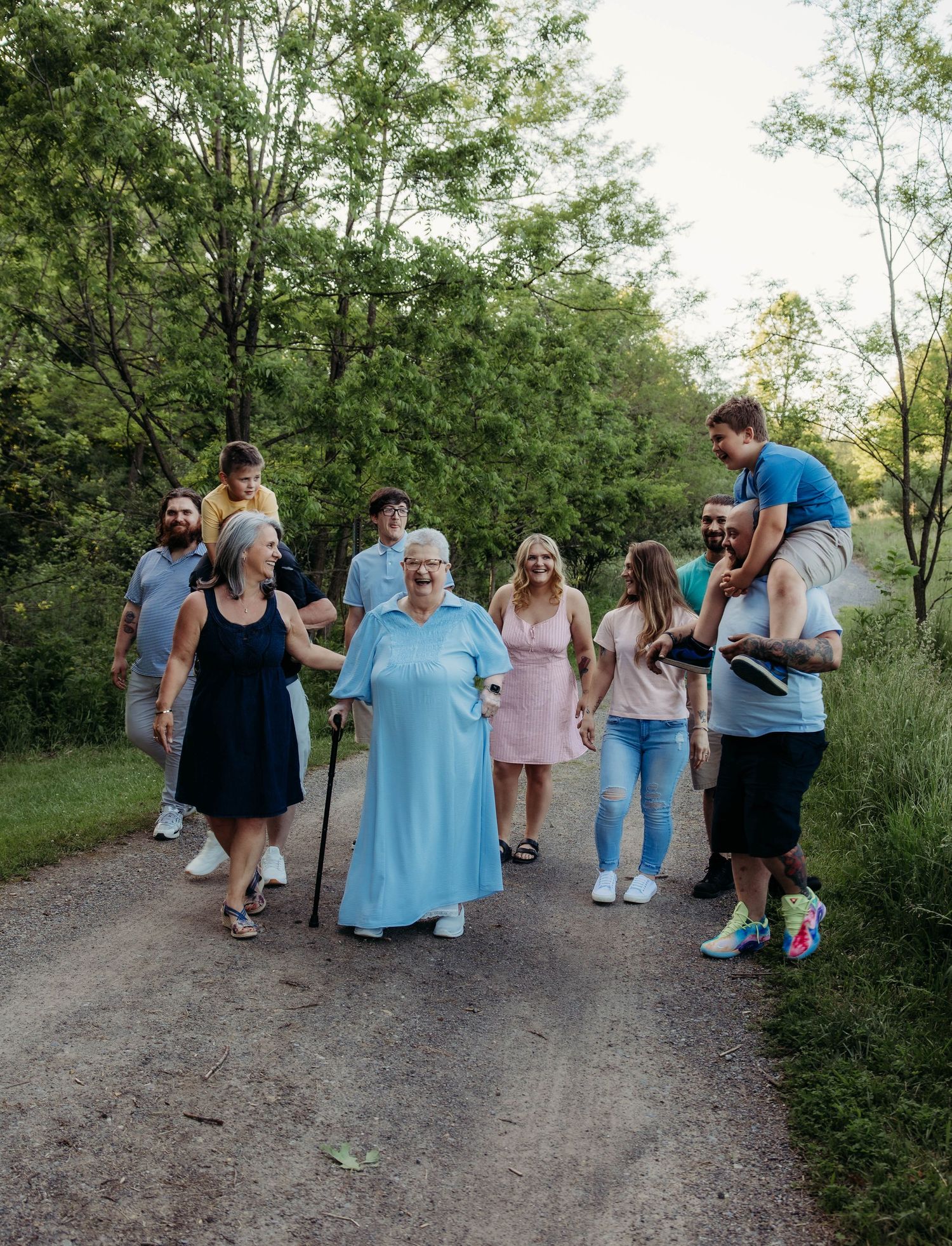 Multi-generational family walking together on a scenic tree-lined gravel path, enjoying an outdoor stroll in nature.