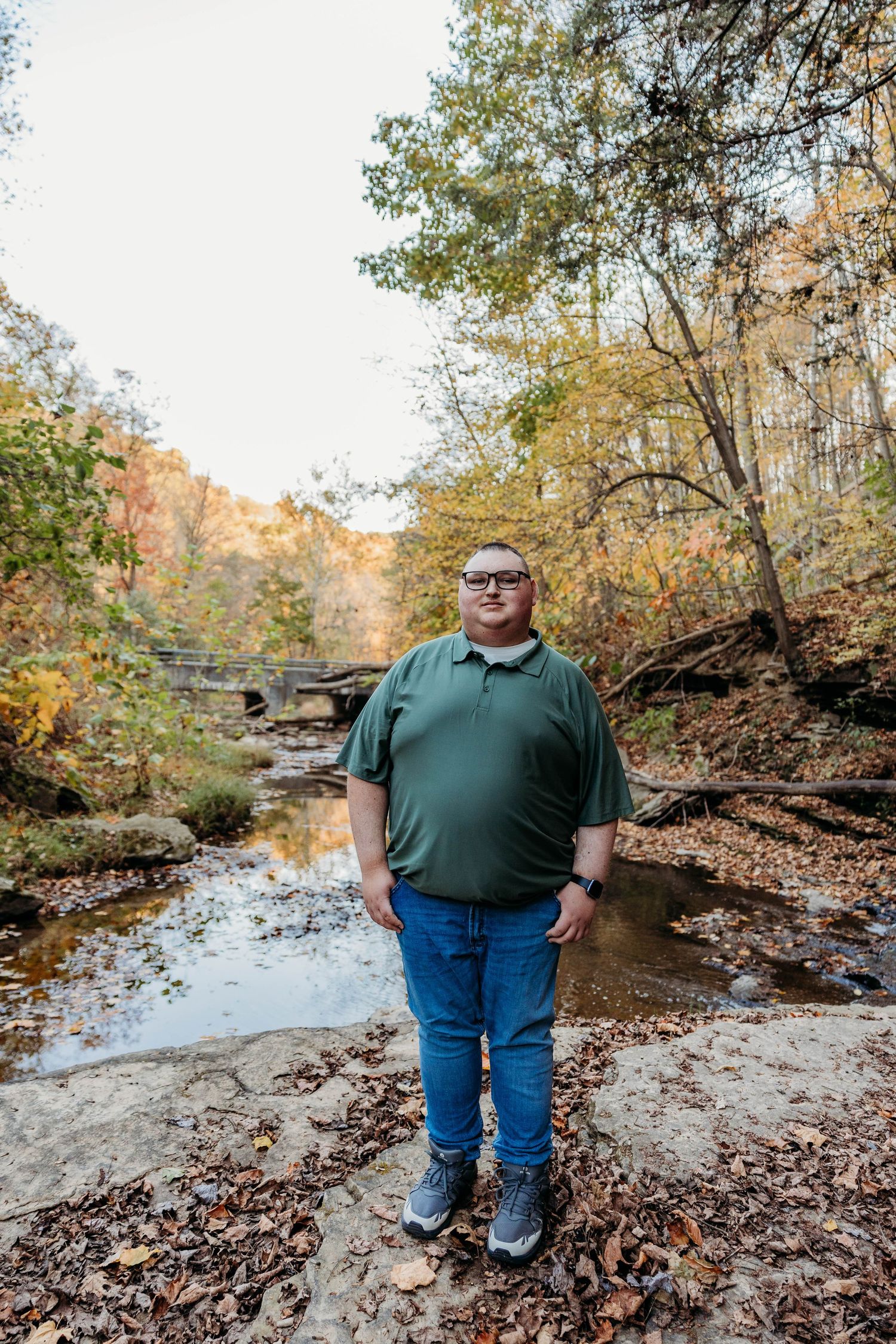 Man in green polo and jeans standing by a rocky creek in an autumn forest with colorful foliage.