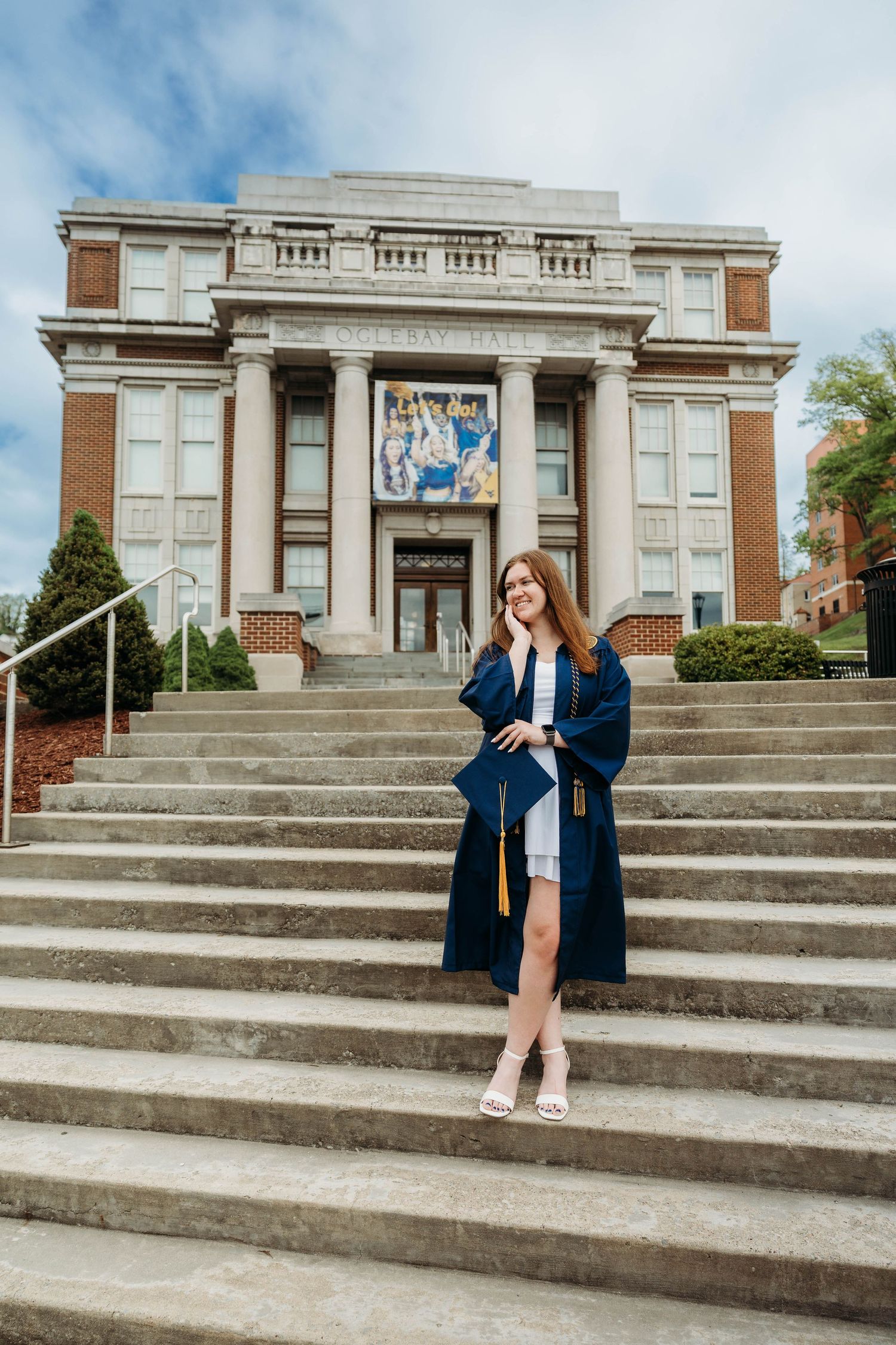 A smiling graduate in navy cap and gown poses on university steps holding a diploma on a cloudy day.