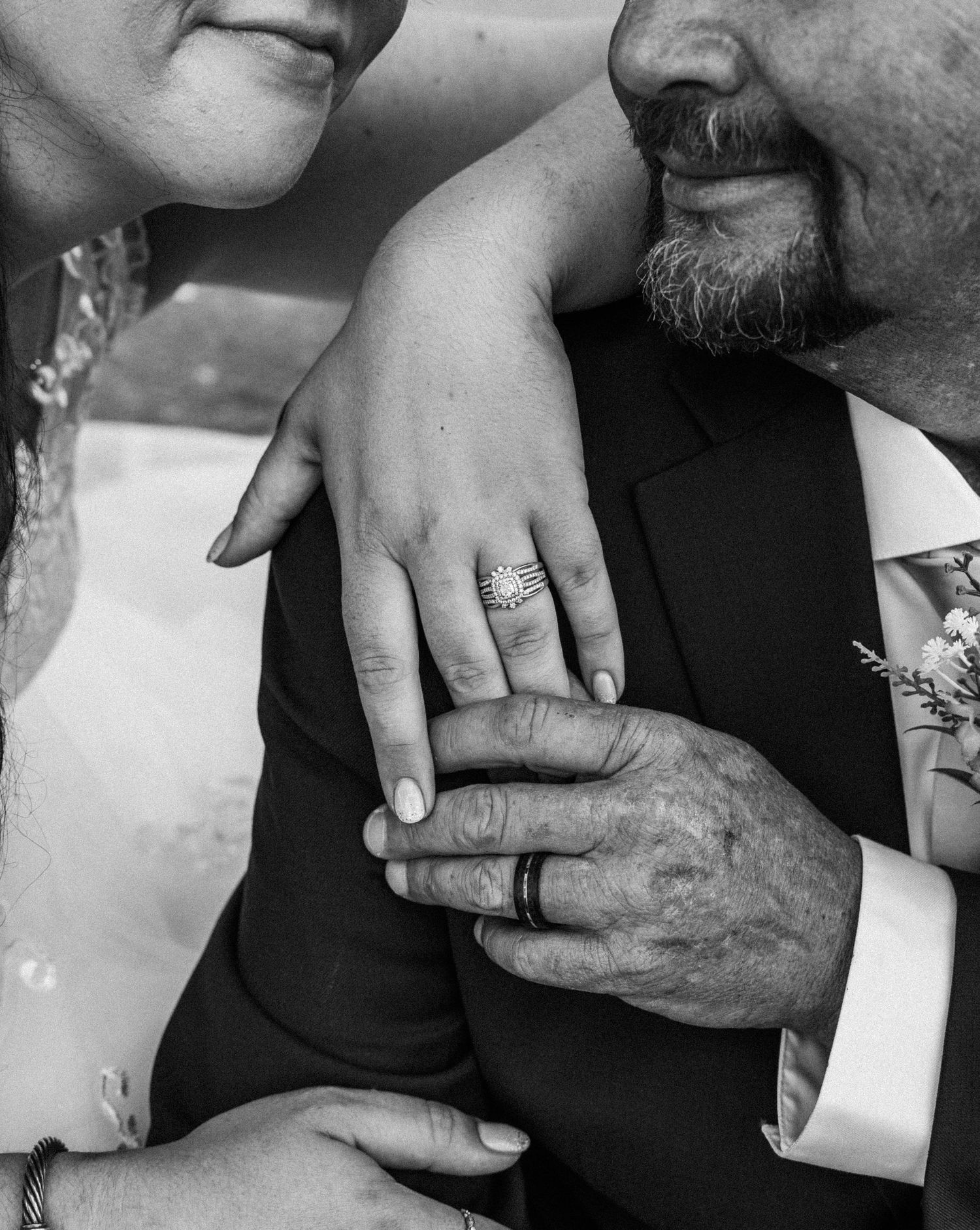 Black and white wedding photo of bride and groom holding hands, showcasing wedding rings, groom in suit with boutonniere.