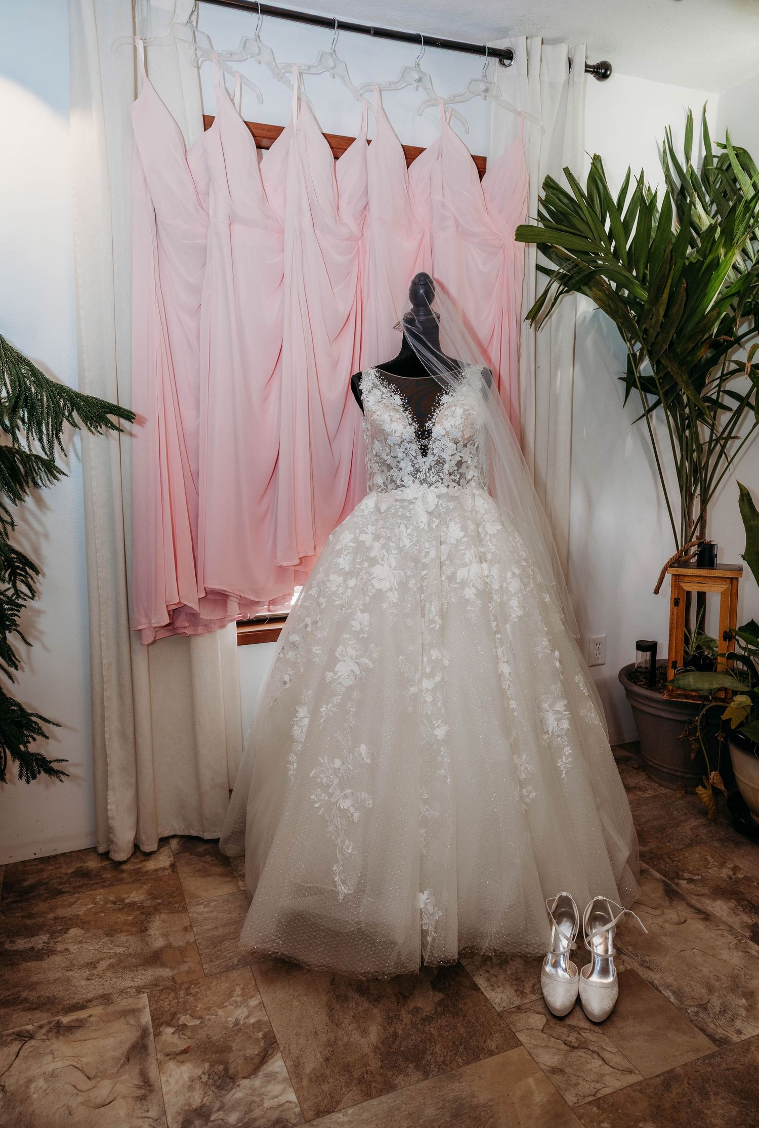 Elegant lace ballgown wedding dress on mannequin with veil, white heels, and pink bridesmaid dresses hanging in background.
