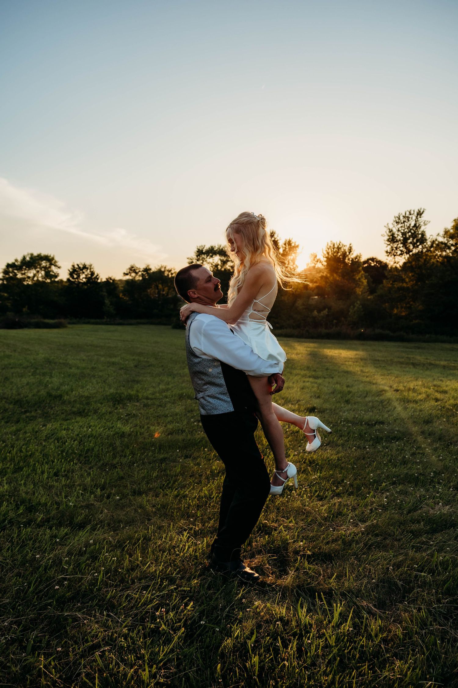 Groom lifting bride in white dress at golden sunset in lush green field, romantic wedding photo outdoors.