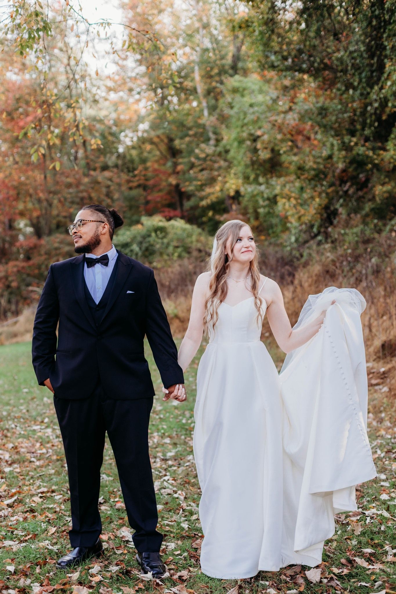 Autumn wedding portrait of couple walking through fallen leaves in formal attire.